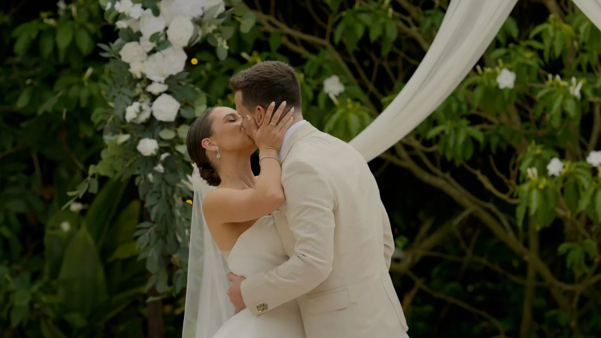 A bride and groom sharing a kiss during their outdoor wedding ceremony surrounded by greenery and white flowers.