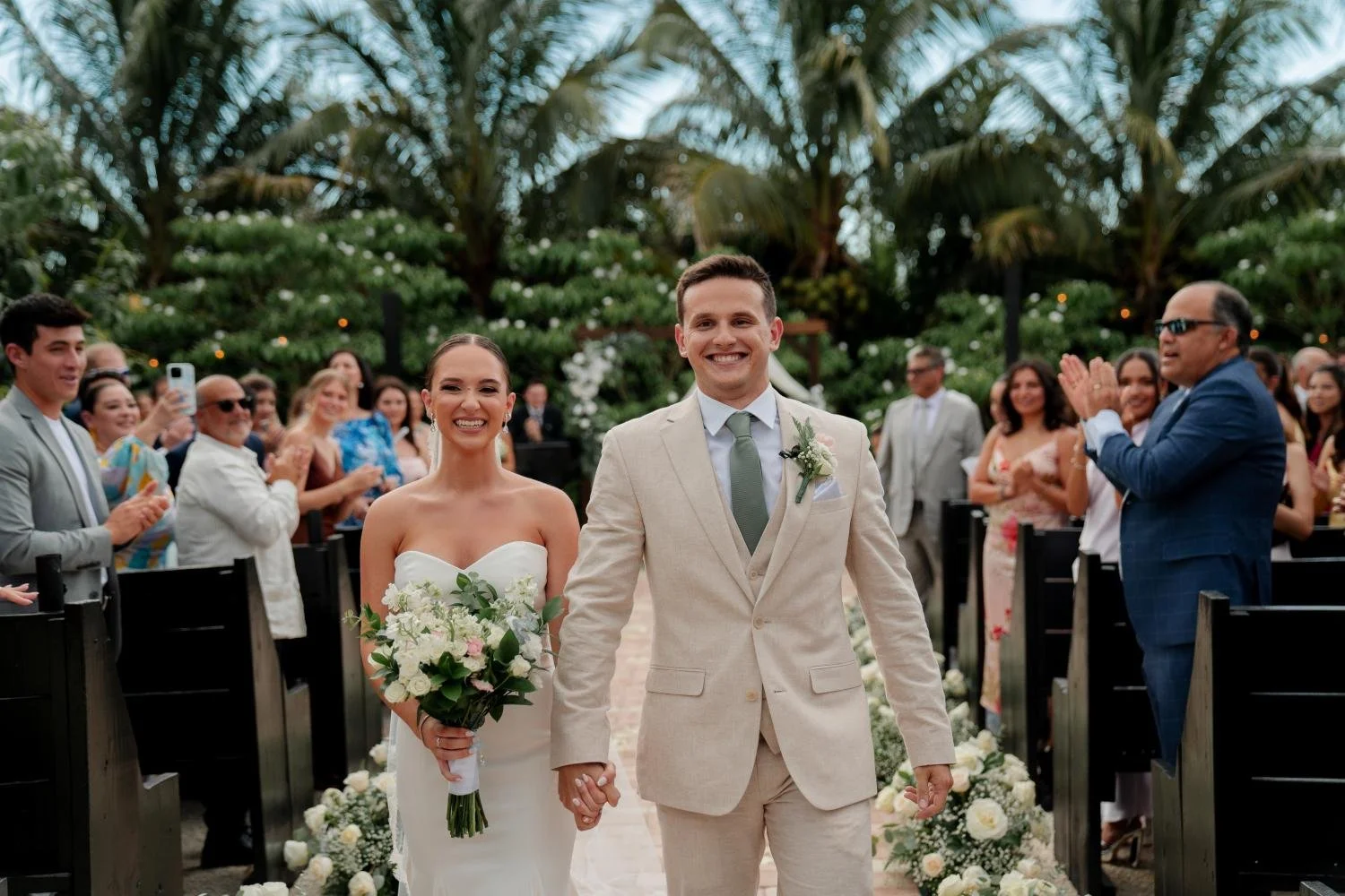 A newlywed couple walking down the aisle at their outdoor wedding ceremony. The bride is holding a bouquet of white flowers, and the groom is wearing a light-colored suit. Guests are smiling and clapping, with some taking photos, in a lush garden set