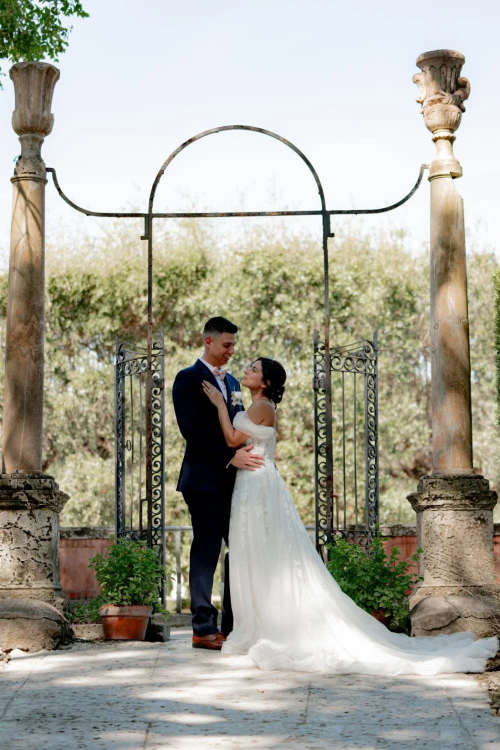 A bride and groom standing under an ornate metal archway on their wedding day, embracing each other and smiling, with lush greenery in the background.