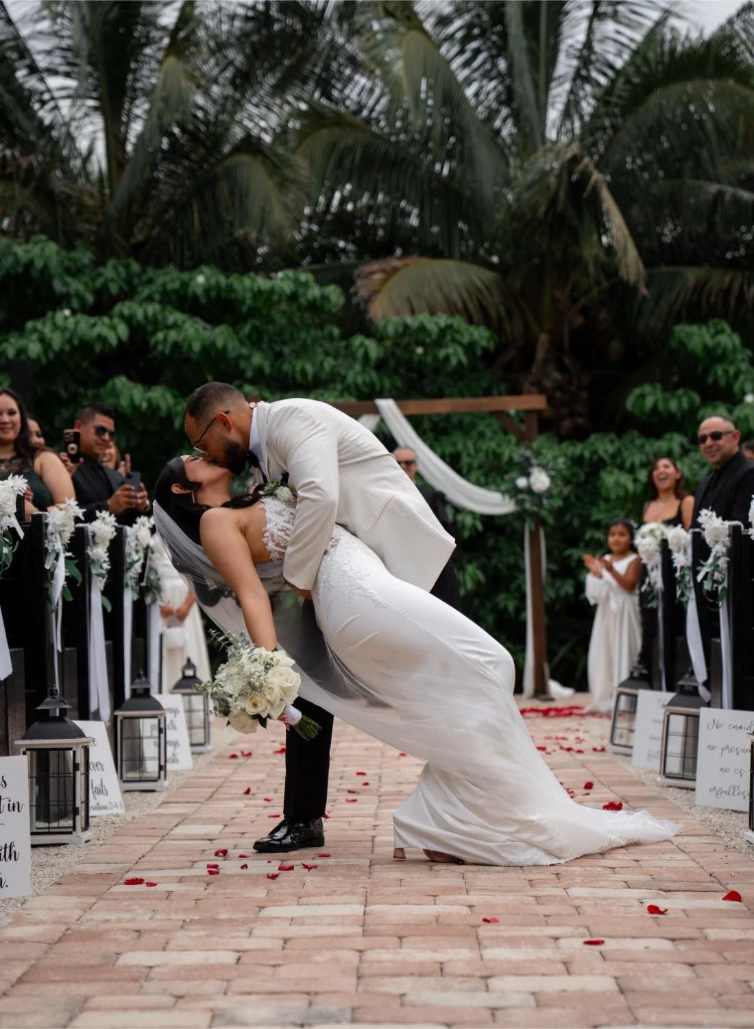 A wedding couple kissing during their outdoor ceremony with guests watching and celebrating in the background, surrounded by lush greenery and decorated with white flowers.