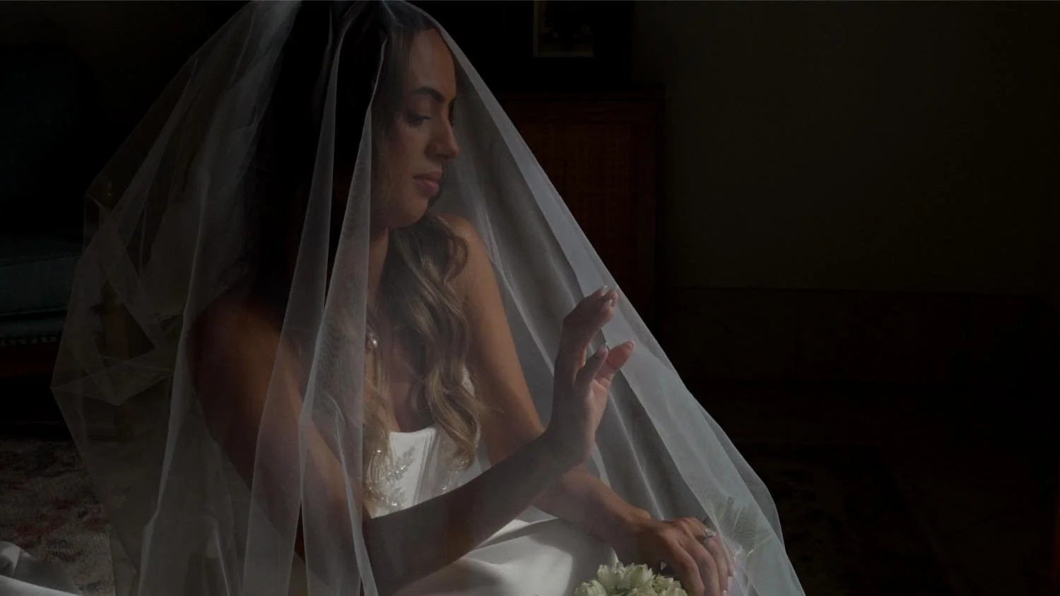 Woman in wedding dress sitting on the floor with a veil over her head, touching her wedding ring with closed eyes in a dimly lit room.