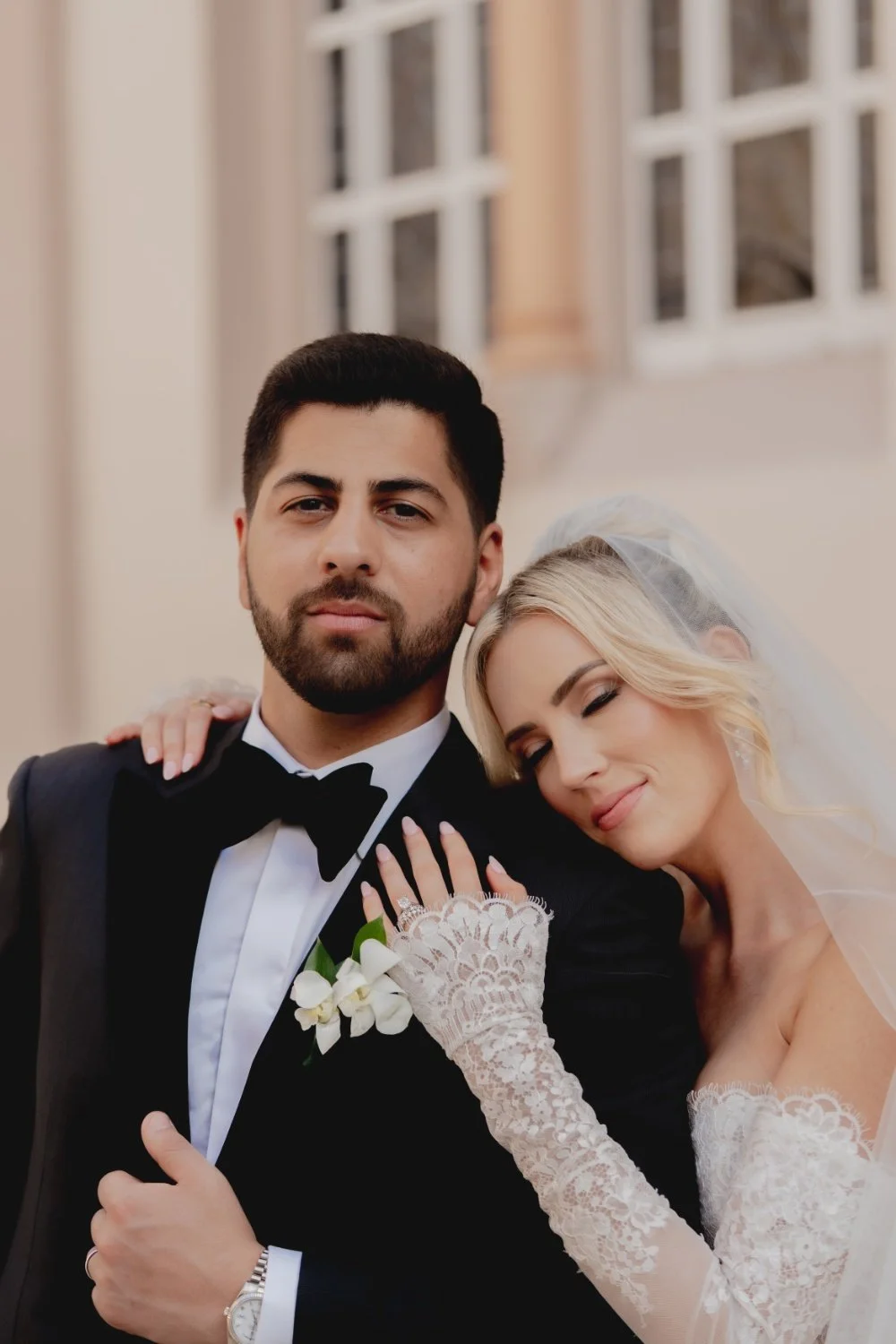 A bride with blonde hair and a groom with dark hair and beard, dressed in a tuxedo, standing close together outdoors near a building with large windows, during wedding.