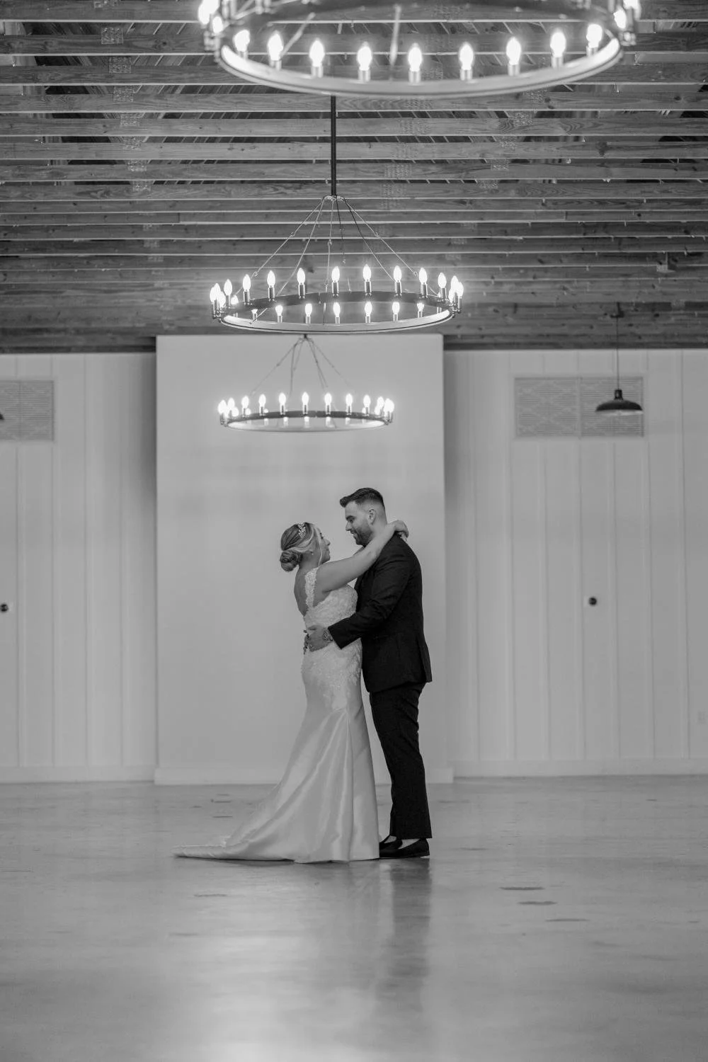 A bride and groom dancing in a wedding reception hall, under hanging chandeliers, with a plain wall background.