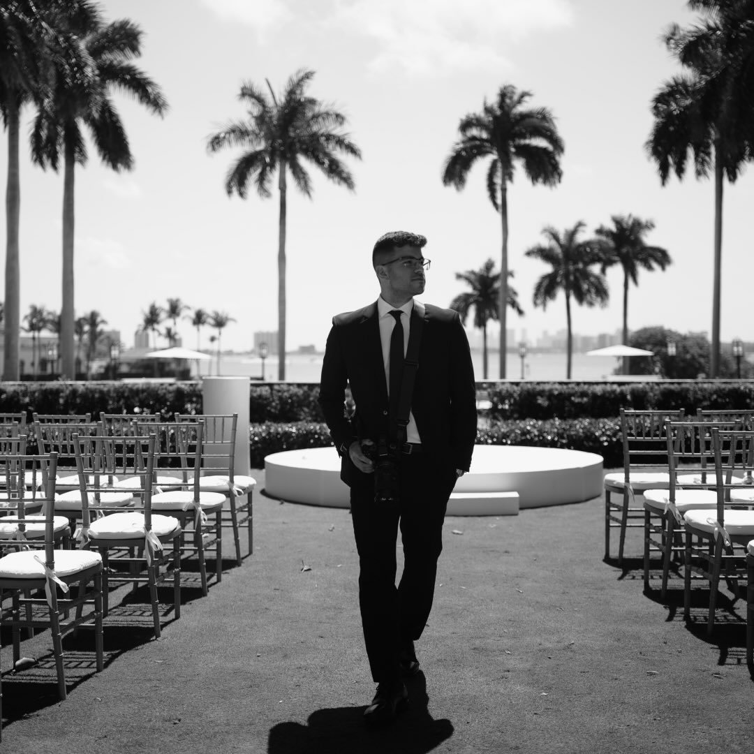 A man in a suit walks through an outdoor area with palm trees, chairs, and a circular platform, with a city skyline in the background, in black and white.