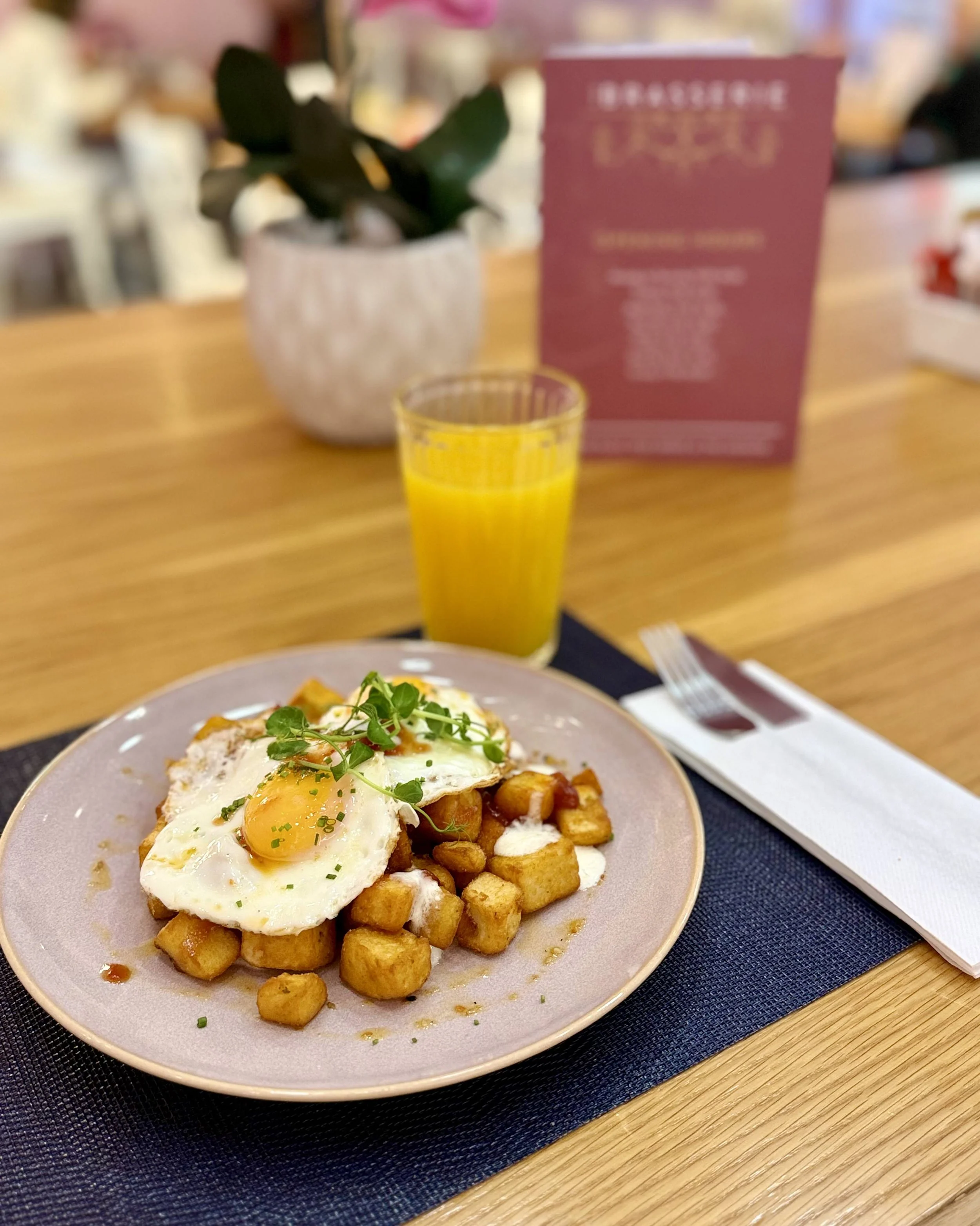 A plate of breakfast food with baked potatoes, a fried egg with a runny yolk, and garnished with green herbs, on a pink plate on a table with a fork, napkin, and glass of orange juice in the background.