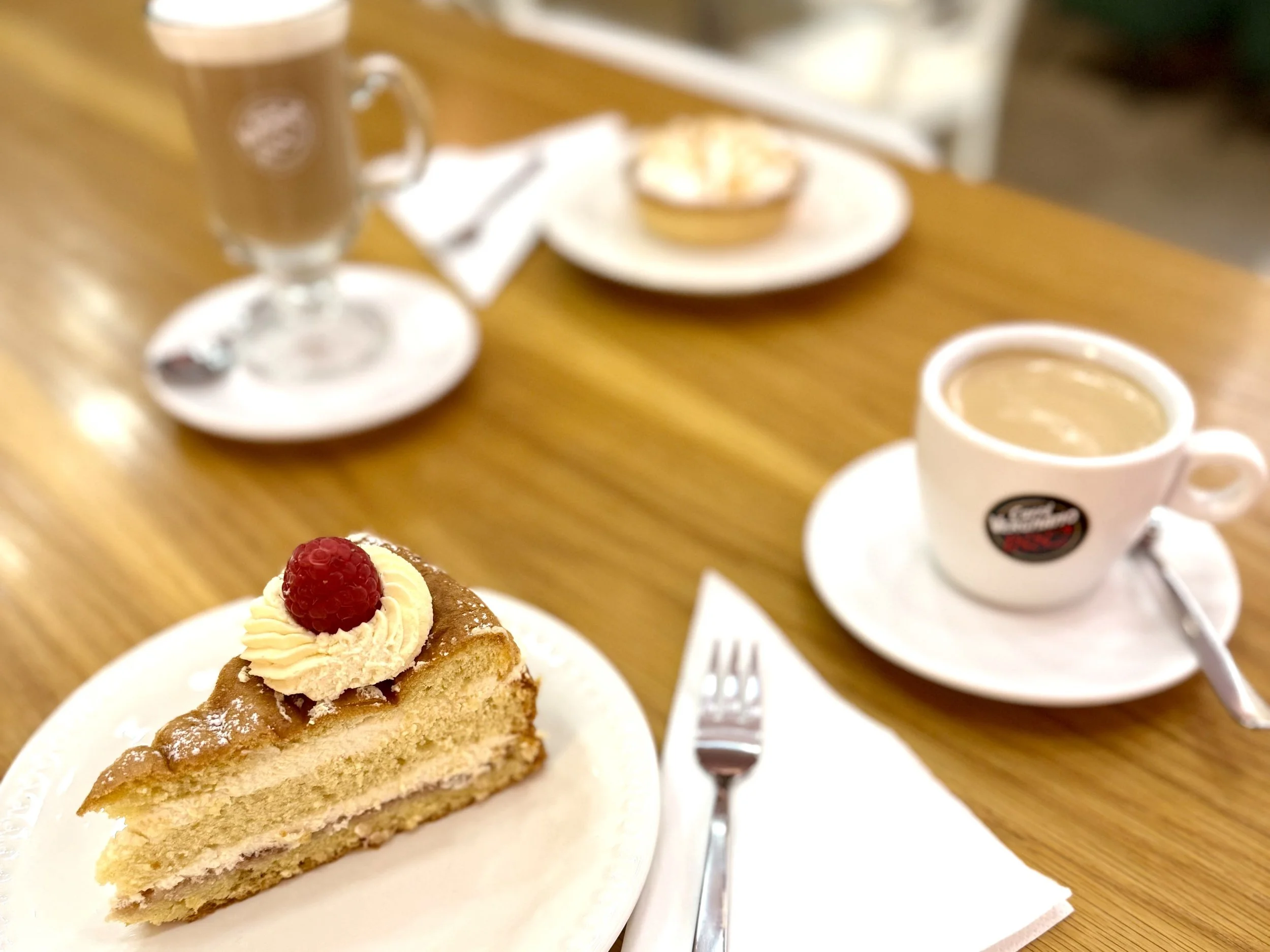 A slice of cake topped with a dollop of cream and a raspberry, served on a white plate. In the background, there is a cup of coffee or tea, a plate with more baked goods, and a mug on a wooden table.
