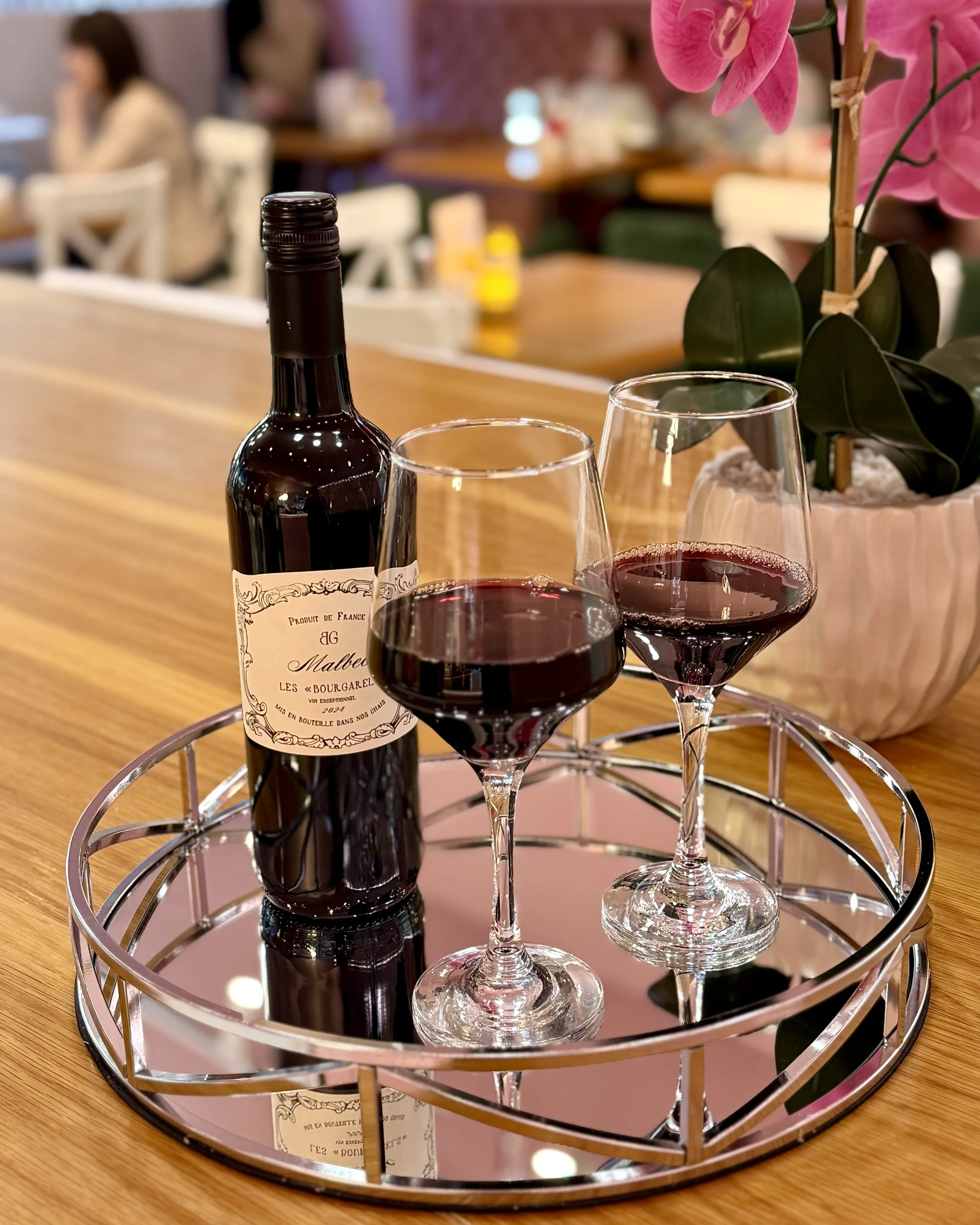 A bottle of red wine and two glasses filled with red wine on a silver tray, with pink orchids in a pot in the background, on a wooden table in a restaurant.