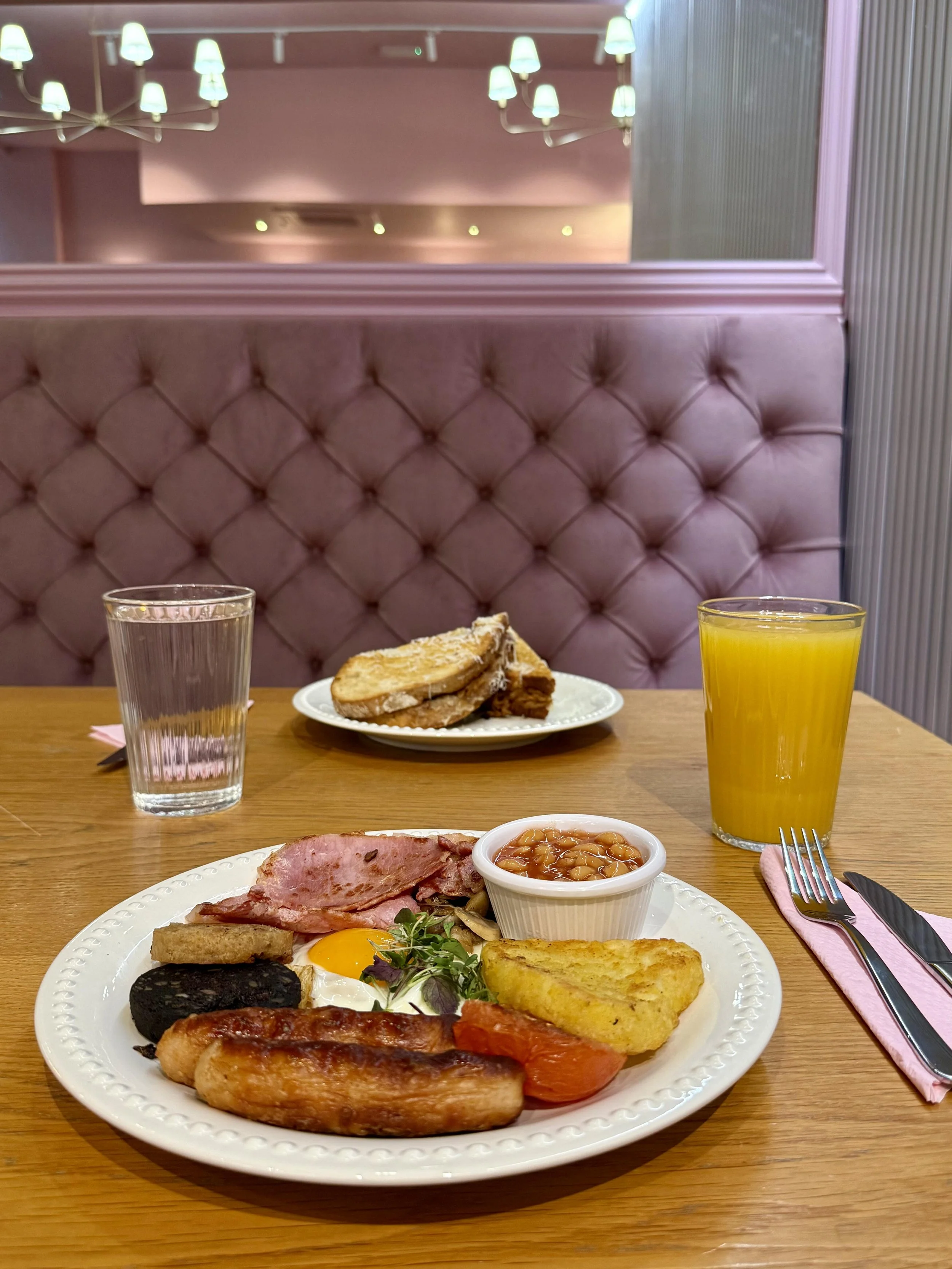 A breakfast meal includes a plate with bacon, toast, an egg, sausage, baked beans, hash browns, and grilled tomato, placed on a wooden table. There is a glass of water, a glass of orange juice, a fork, and a knife with a pink napkin.