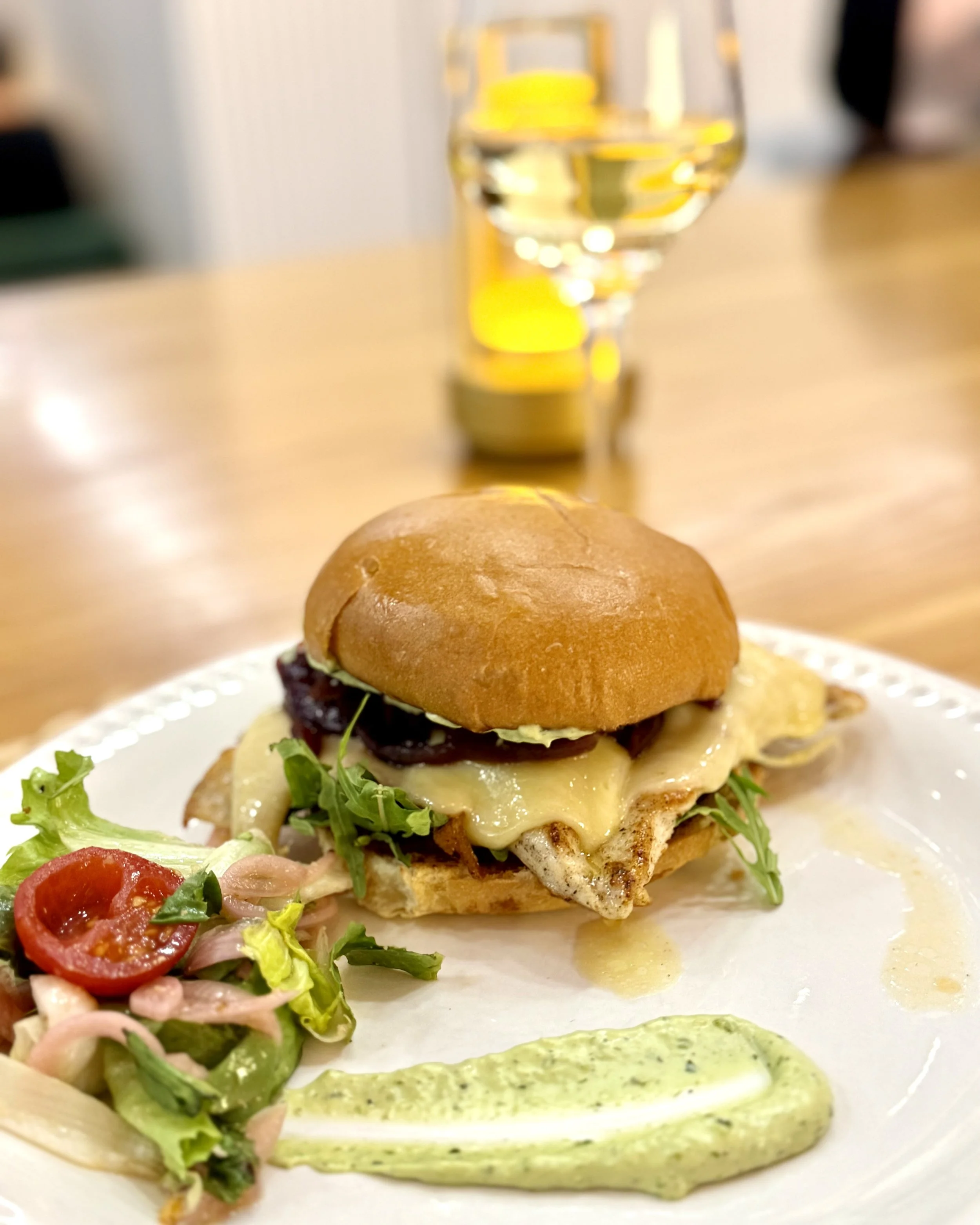 A close-up of a burger with lettuce, cheese, and cooked chicken inside a bun, with a side salad and a serving of green sauce on the plate.