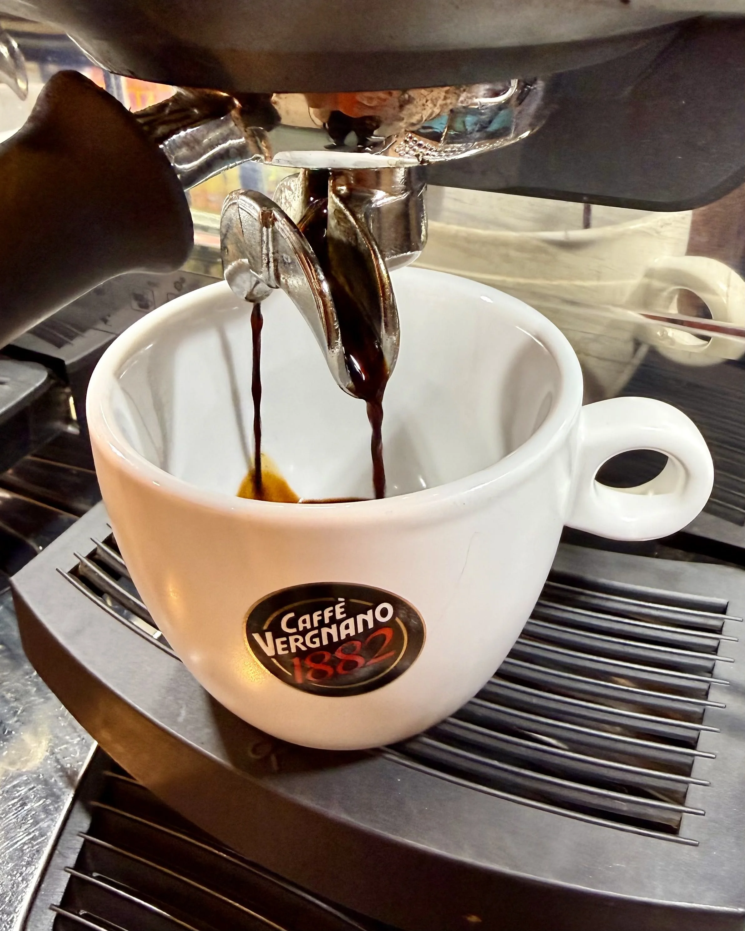 A white coffee mug with the logo 'Caffè Vergnano 1882' is placed on a black stovetop. Hot black coffee is being poured into the mug from a metallic espresso machine spout.
