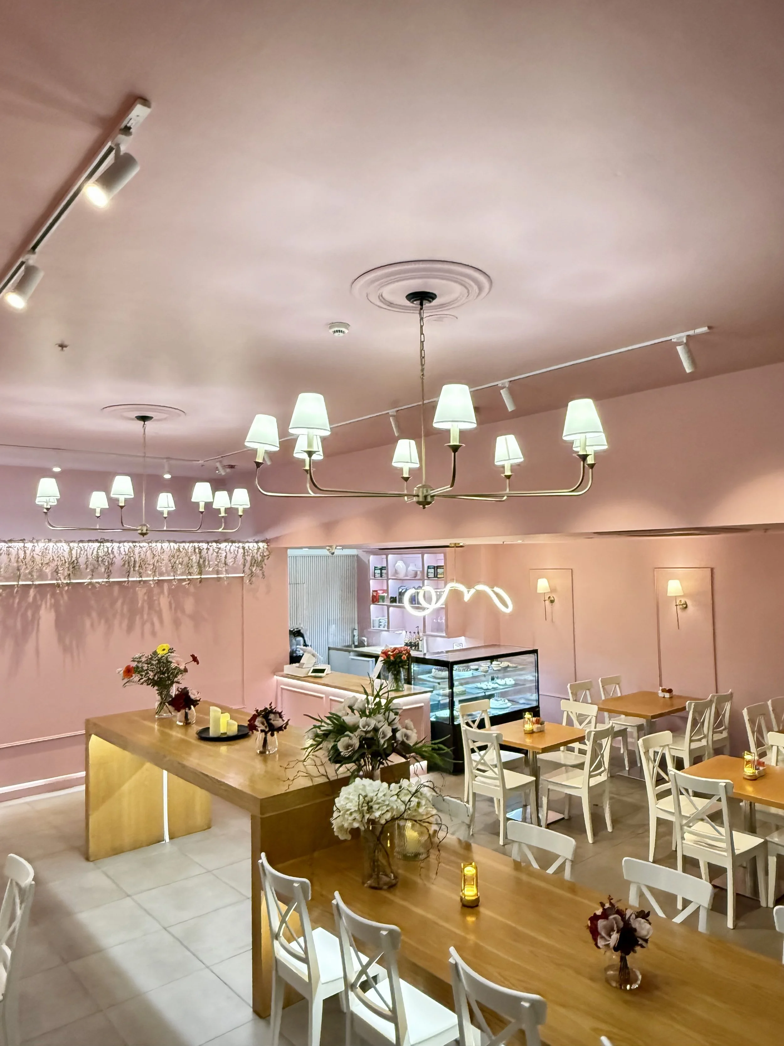 A cozy cafe interior with pink walls, white chairs, and wooden tables decorated with flowers and candles. Overhead, there are modern chandeliers and track lighting.