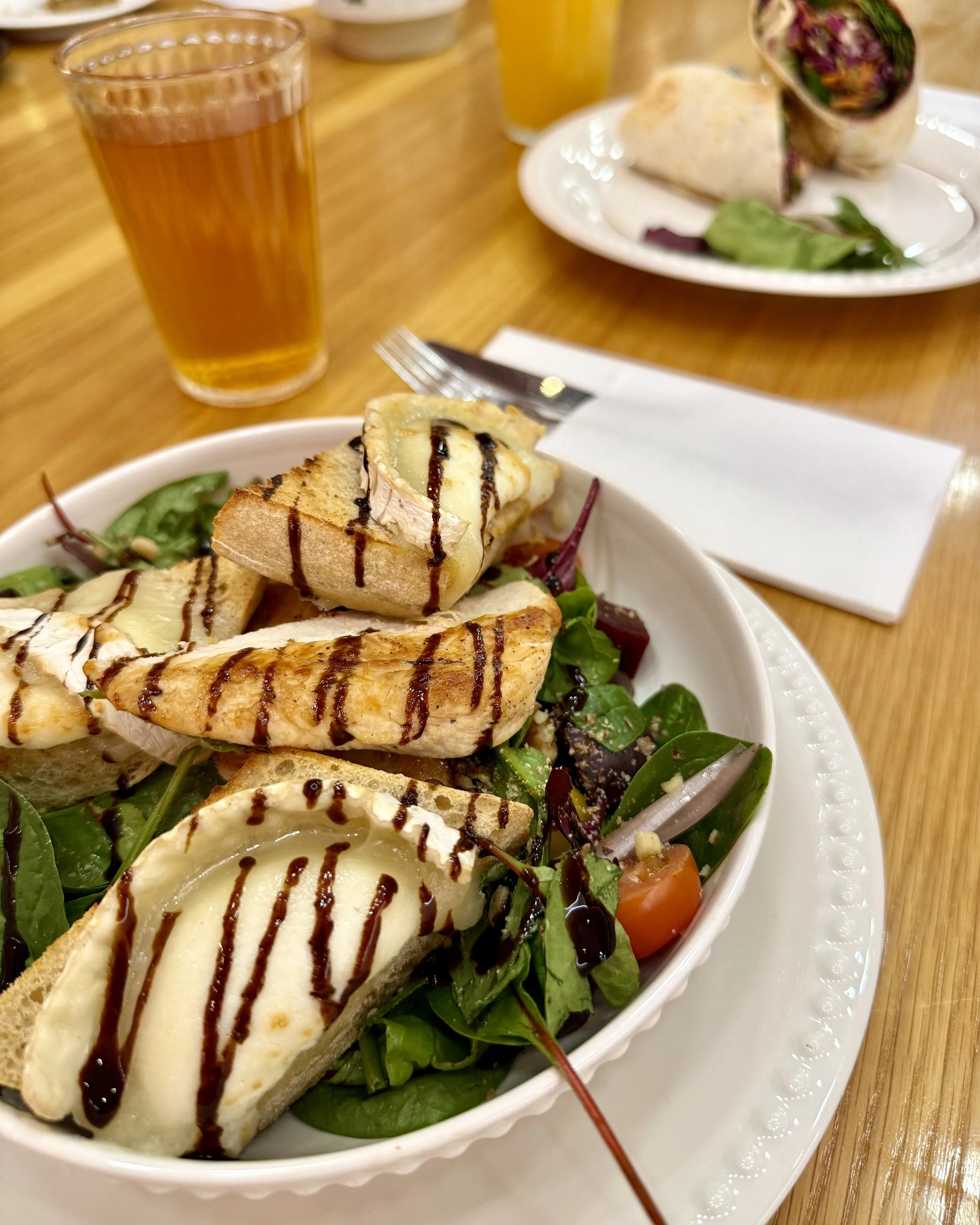 A bowl of salad with grilled chicken, slices of cheese, tomato, avocado, and salad greens drizzled with balsamic glaze, on a white plate on a wooden table.