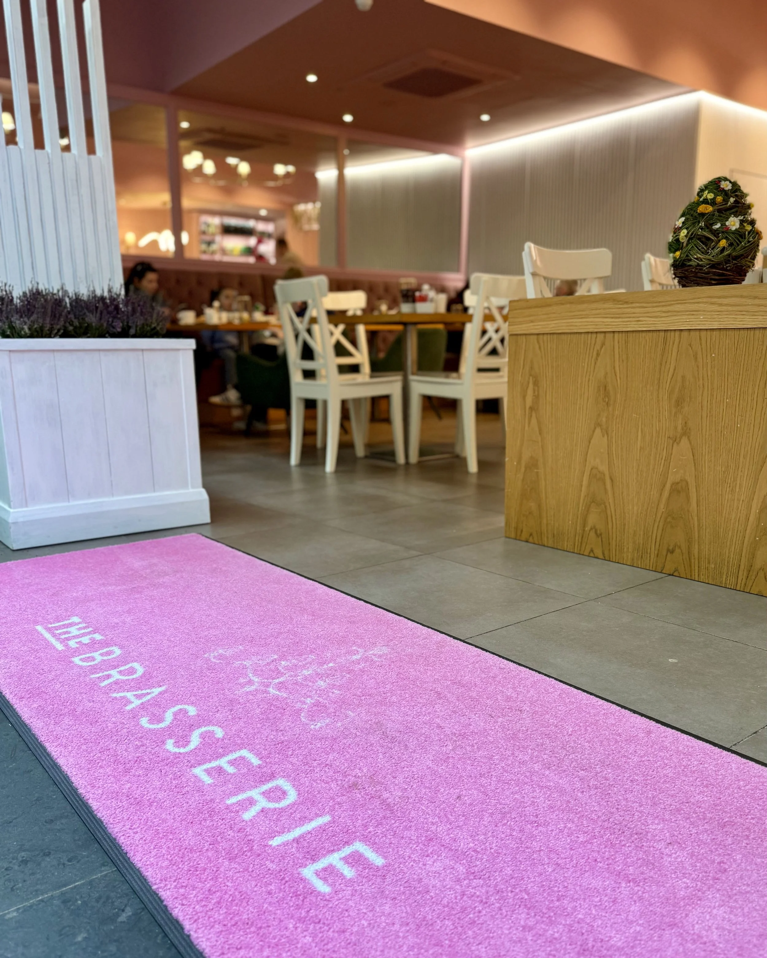 Indoor scene with a pink floor mat that has white text, wooden furniture, decorative flowers, and people dining in the background.