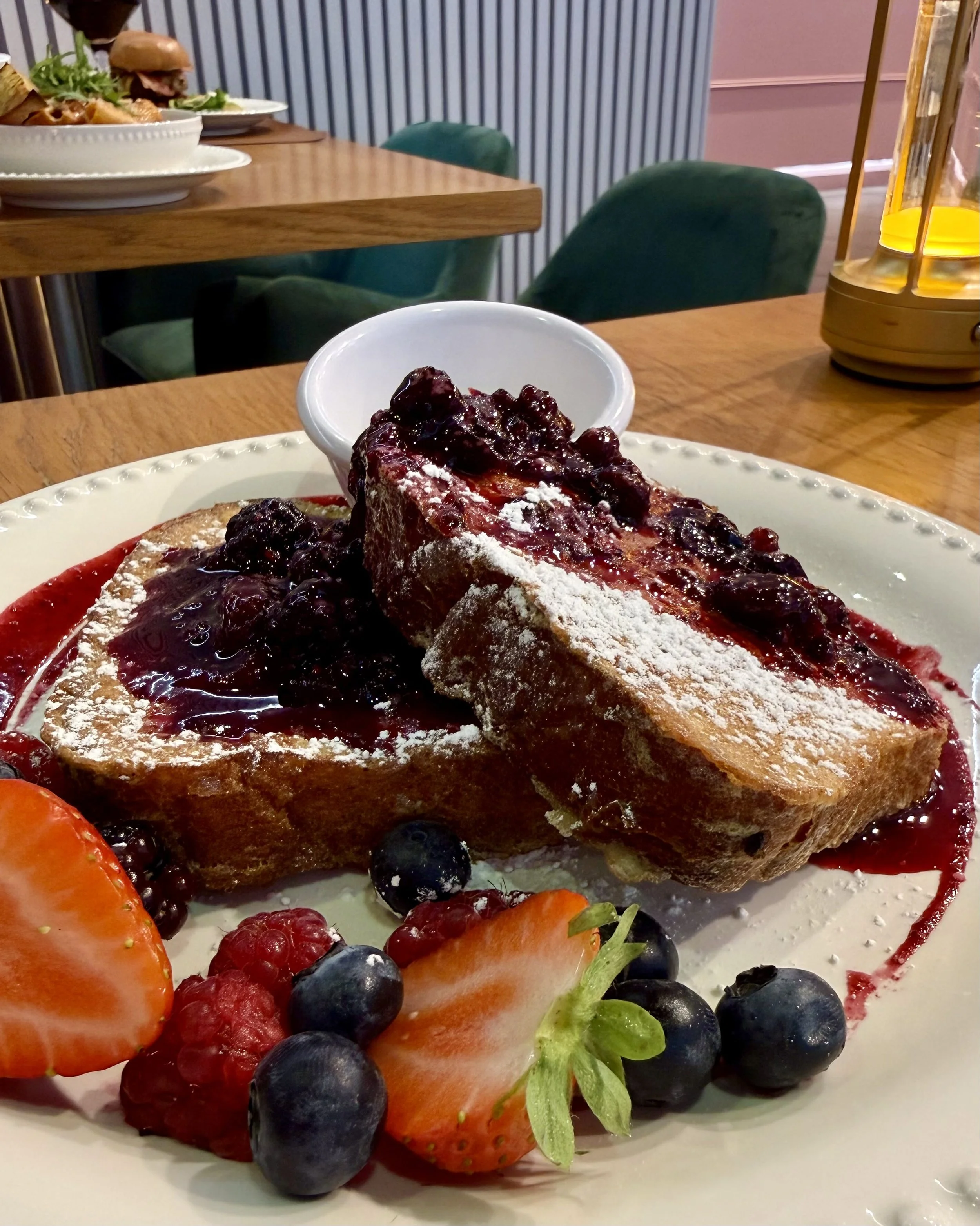 Close-up of a slice of French toast topped with blueberry compote and powdered sugar on a white plate. Fresh strawberries, blueberries, raspberries, and a green leaf garnish are on the side, with a small white bowl nearby. In the background, there are plates with sandwiches and a glass lamp on a wooden table.