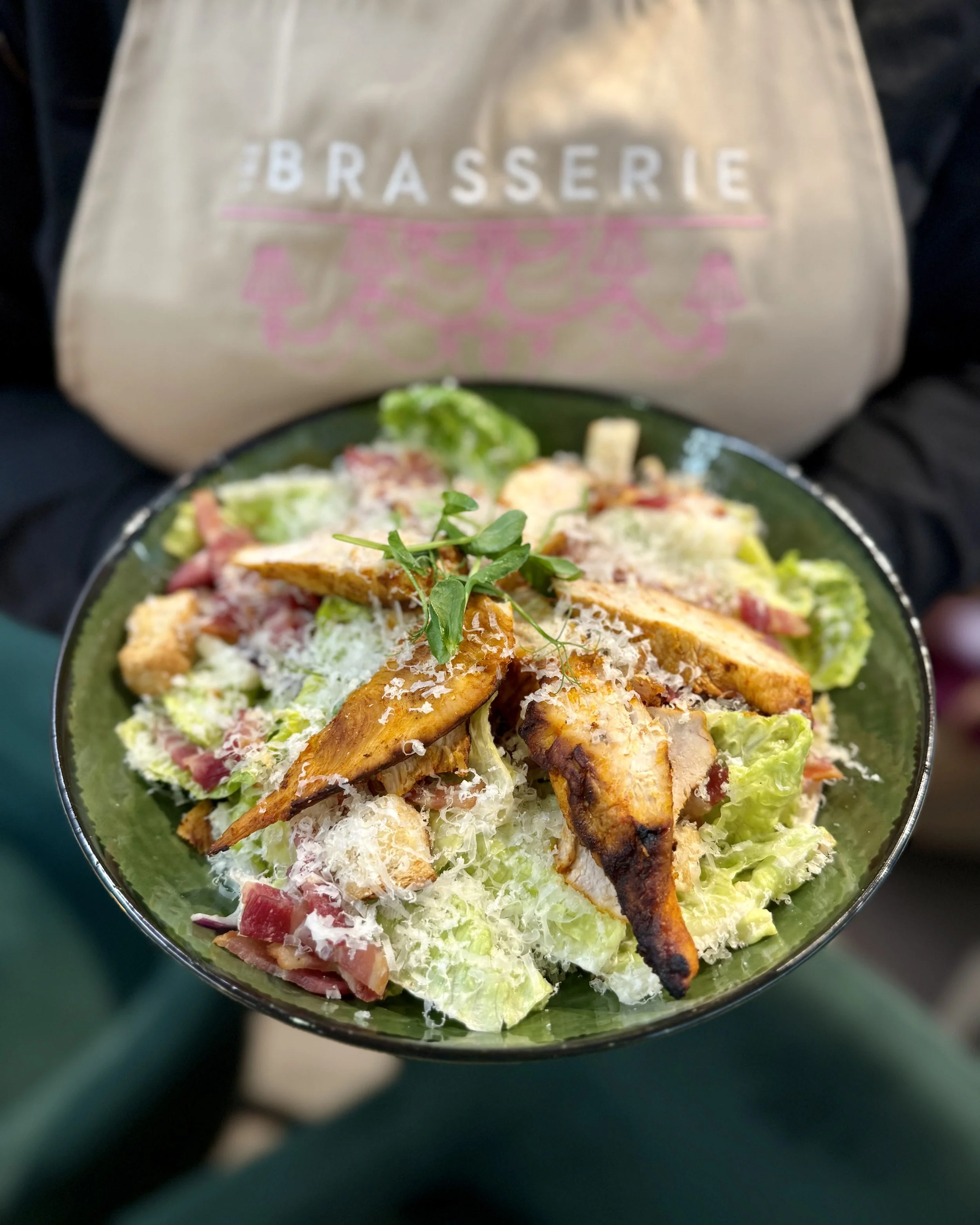A bowl of Caesar salad with grilled chicken strips, garnished with microgreens, shredded cheese, and crispy croutons. In the background, a beige reusable bag with the word 'Brasserie' printed on it.