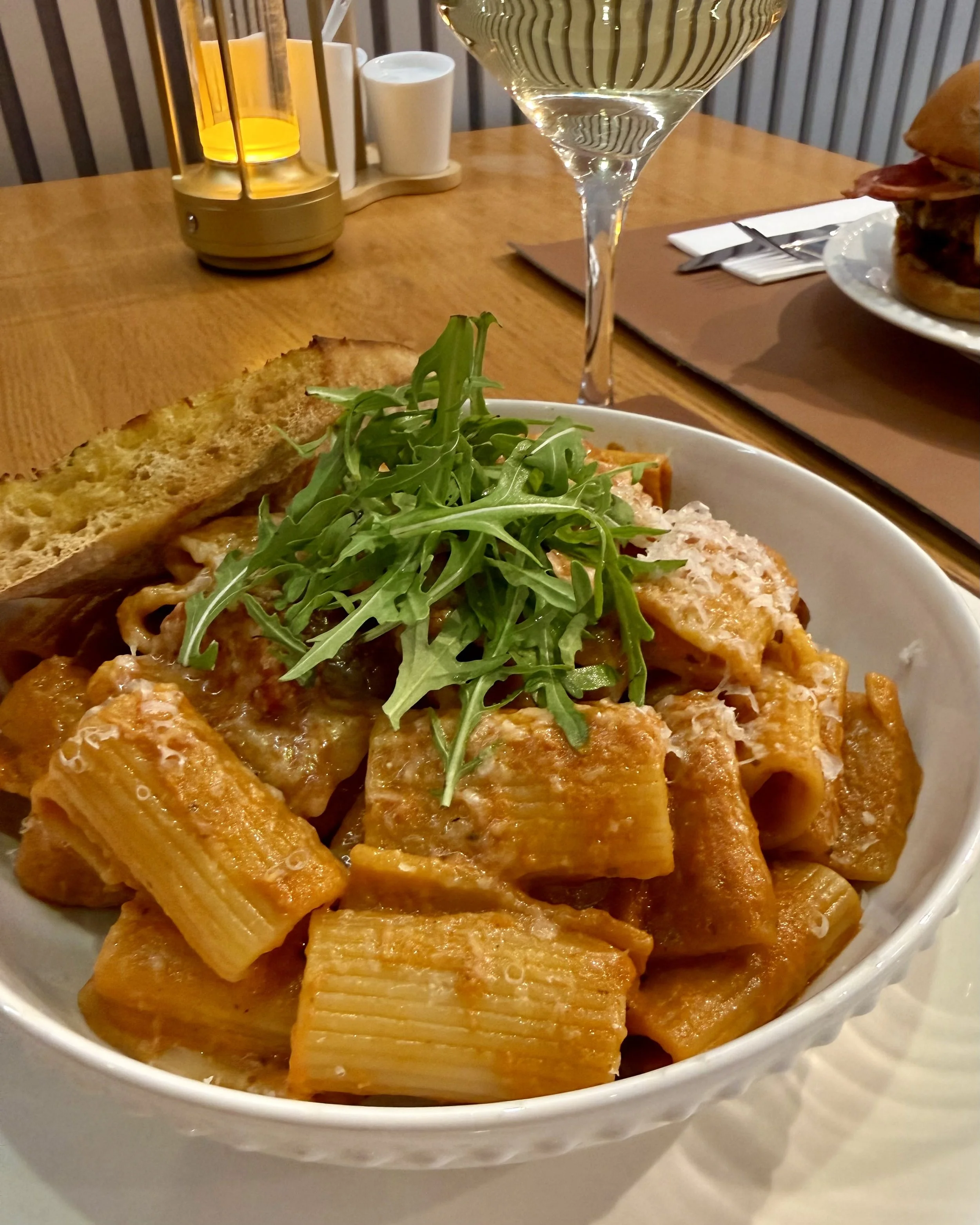 A white bowl filled with fried rectangular food pieces topped with a handful of arugula. In the background, there is a tall glass of white wine, a plate with a sandwich, and a table setting with a knife, fork, and napkin.