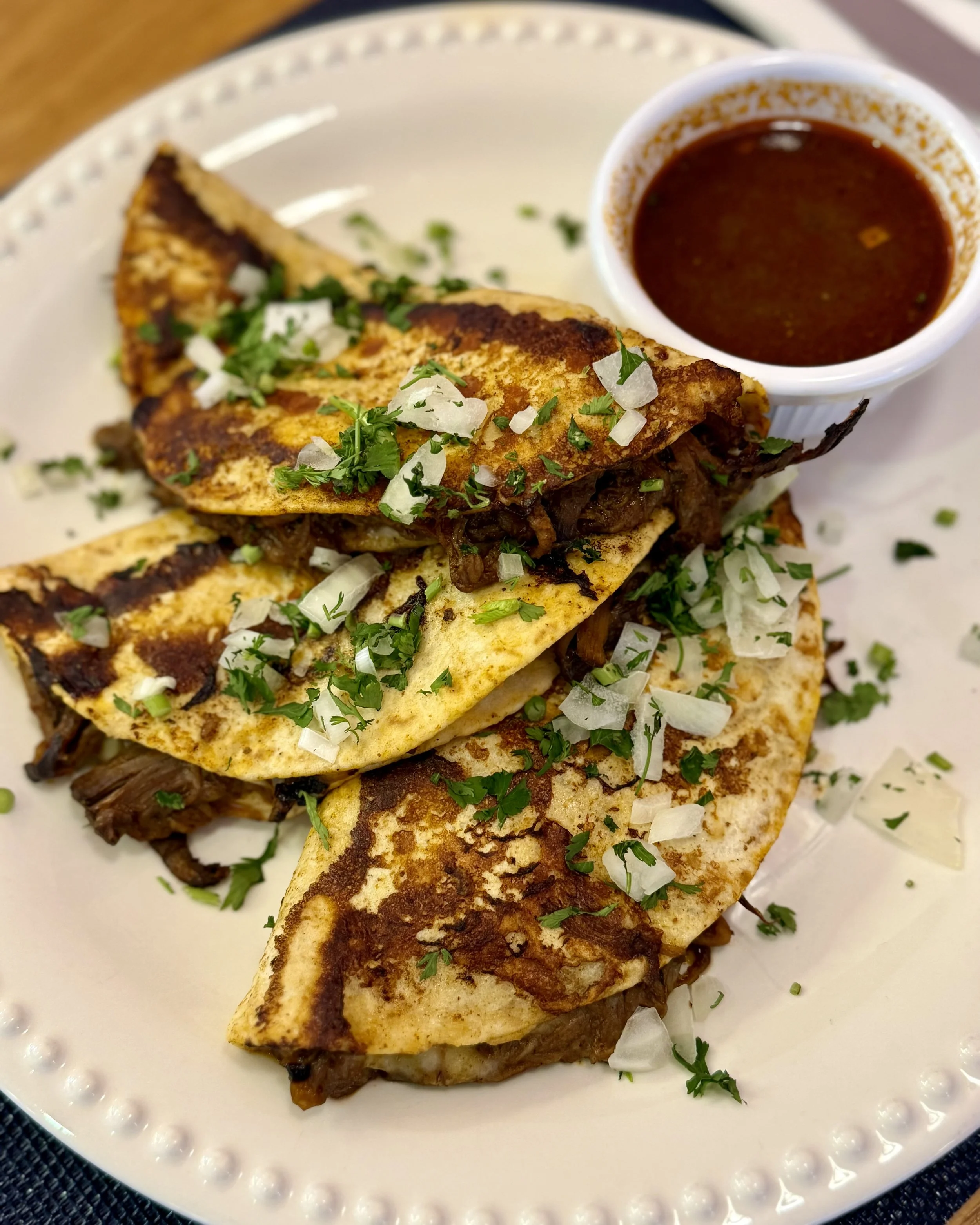 Close-up of tacos on a white plate, garnished with chopped onions and cilantro, accompanied by a cup of dark red salsa.