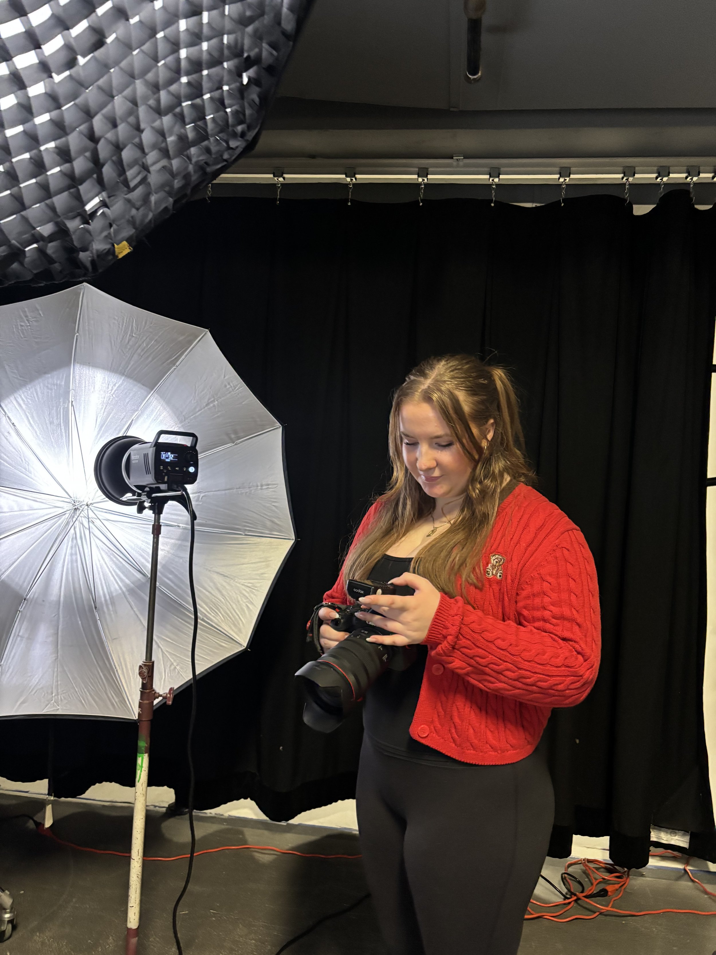 A young woman with long brown hair wearing a red knit sweater is standing in a photo studio, holding a camera and looking down at her phone. There is a large white photographer's umbrella with a light inside it, and a softbox with a black grid pattern on the left side. A black curtain or backdrop is behind her, and various lighting and electrical cords are visible on the floor.