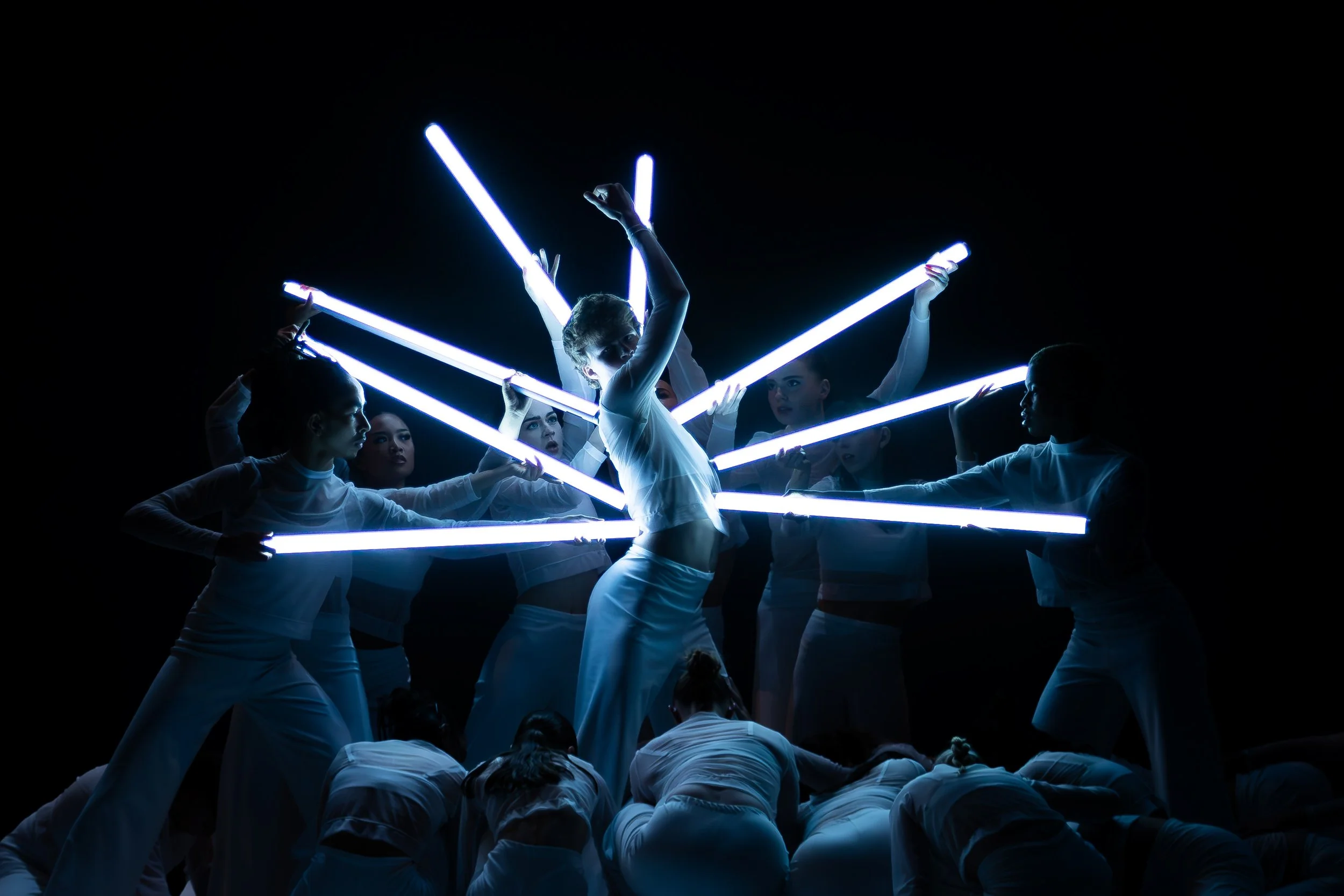 A group of performers in white outfits illuminated by blue neon light sticks, forming a star-like pattern against a black background.
