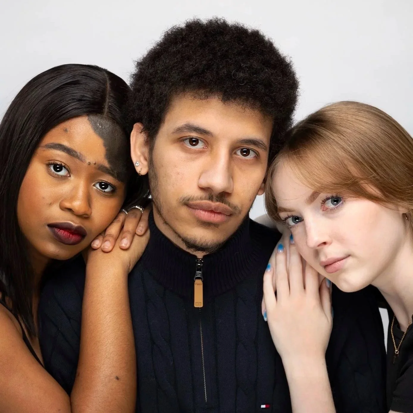 Three young adults closely grouped together, two women and one man, with neutral expressions, against a plain white background.