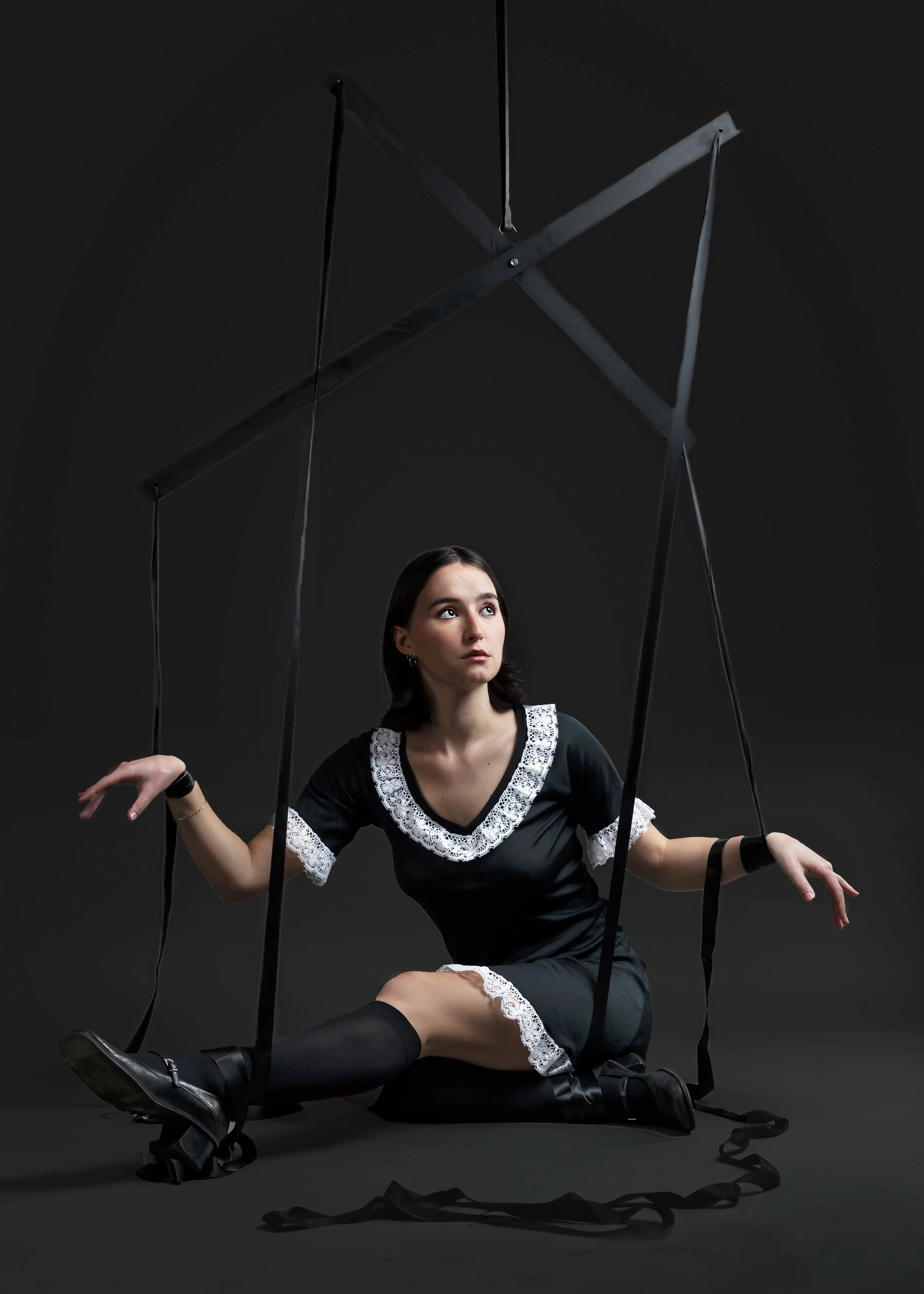 A young woman with dark hair sits on a black swing set against a dark background. She is dressed in a black dress with white lace trim, black knee-high socks, and black shoes. Her arms are extended to her sides, and she gazes upward with a calm expre
