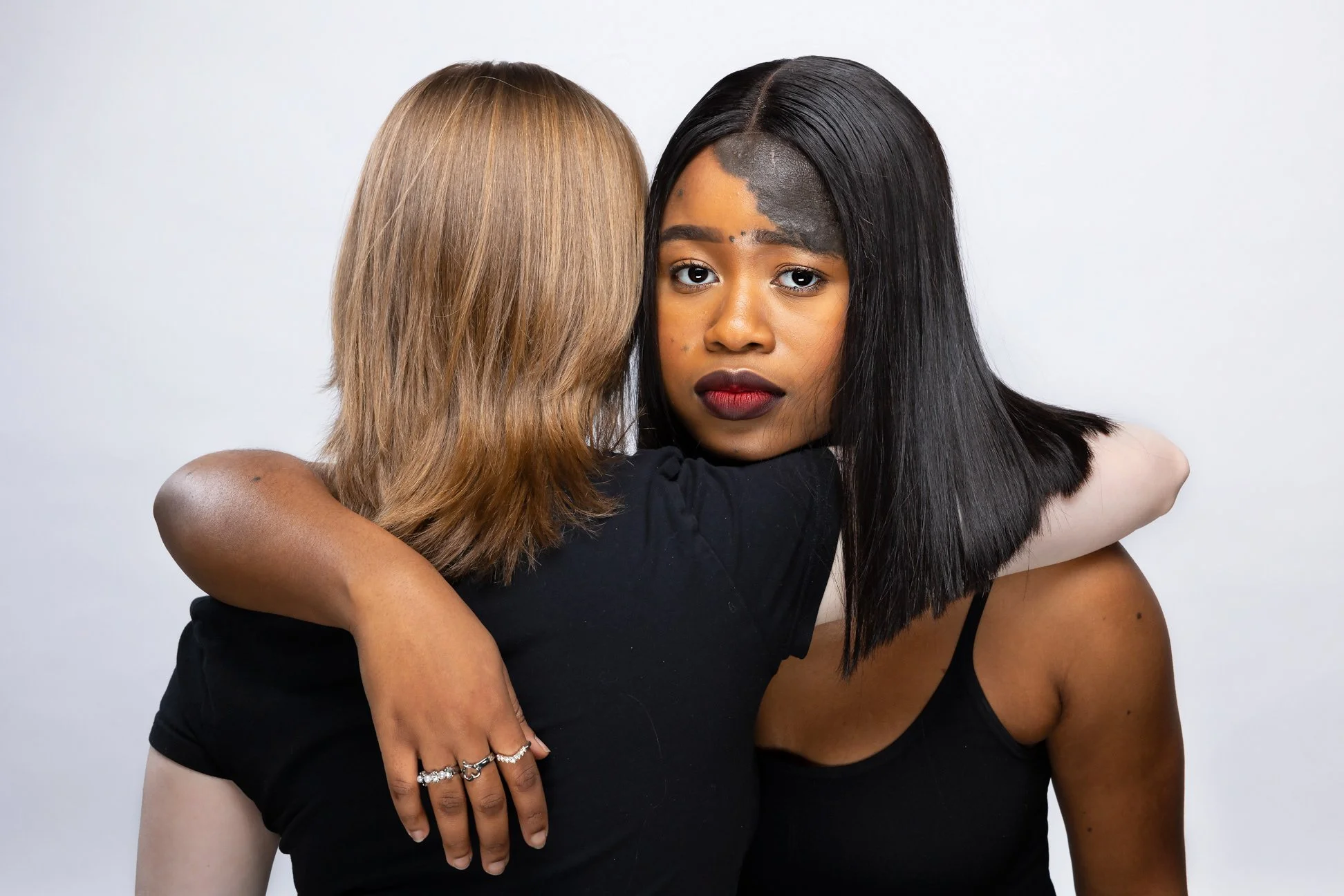Two women hugging each other, one with blonde hair and the other with black hair, both wearing black tops against a plain white background.