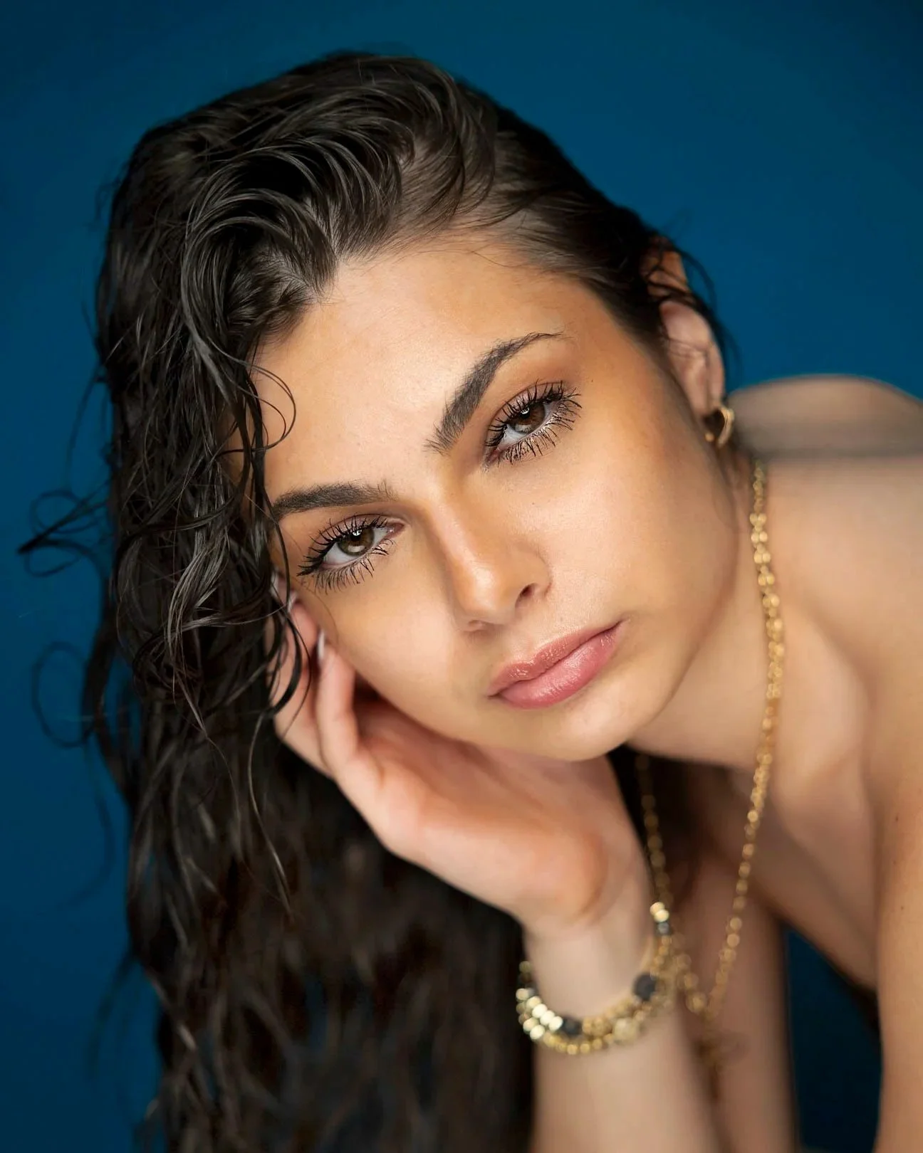 Close-up portrait of a young woman with dark, wet hair, brown eyes, and light makeup, resting her face on her hand, against a blue background.