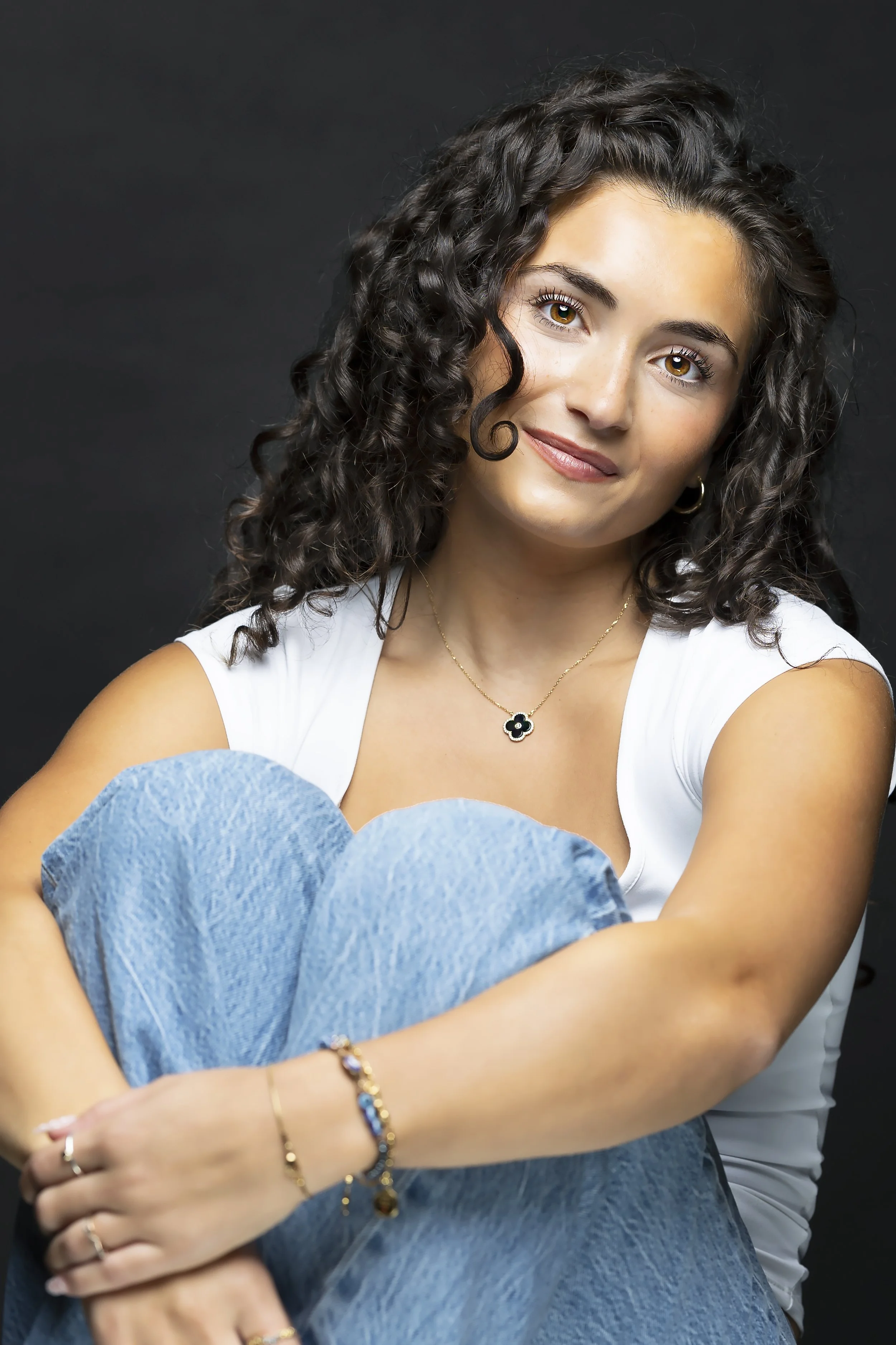 A young woman with curly brown hair, hazel eyes, and a gentle smile, sitting with her arms crossed over her knees, wearing jewelry and a white top against a dark background. Creative professional headshot for an Interior designer.
