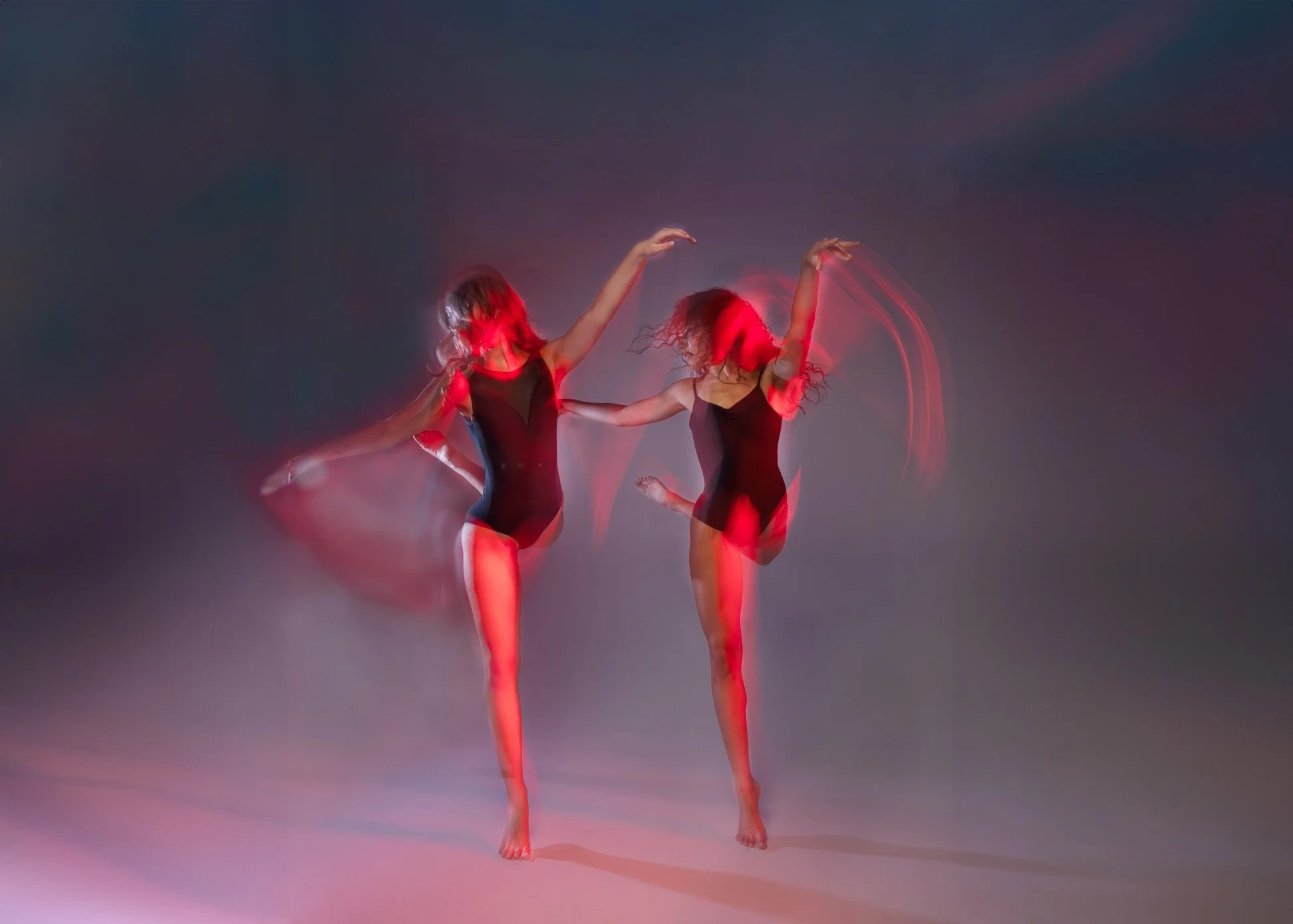 Two dancers are performing in a dark space, wearing black leotards, with pink and red lighting creating motion effects around them.
