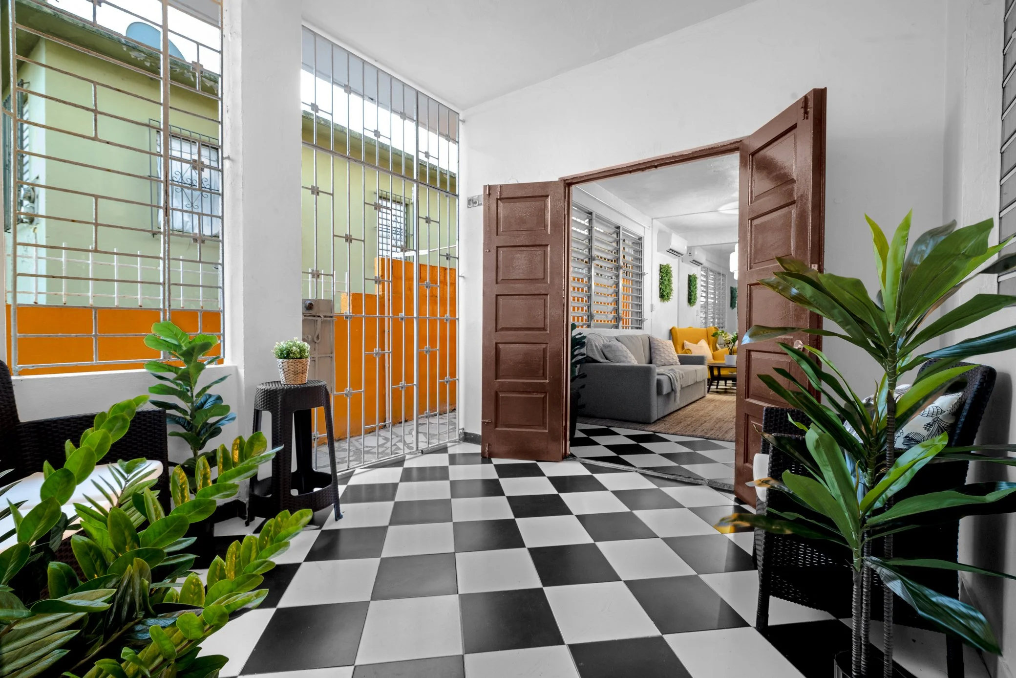 Interior of a house with black and white checkered floor, green plants, black chairs, open wooden double door leading to a living room with gray sofas, yellow pillows, and decorative green plants, and windows with metal bars.