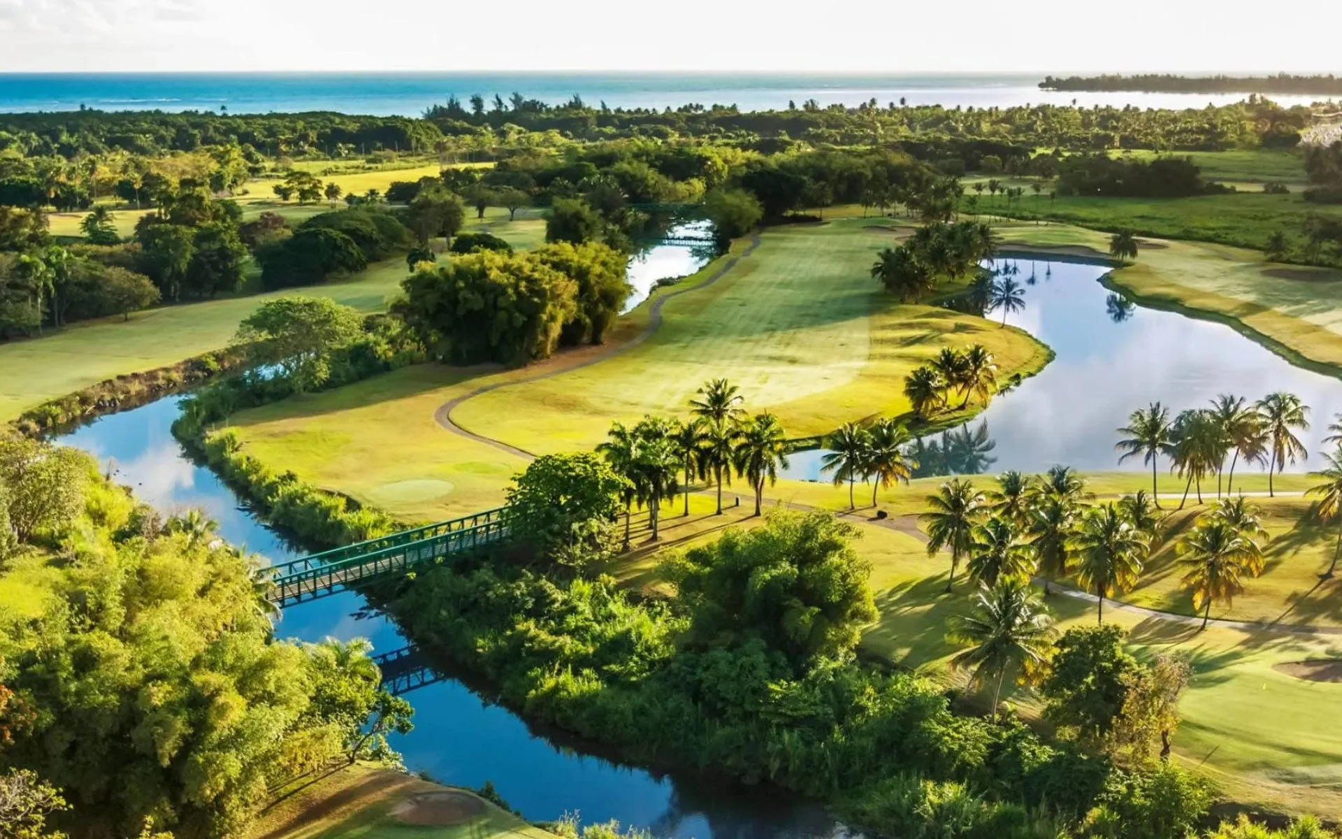 A scenic aerial view of a golf course with ponds, lush green trees, and tropical palm trees near a coastline.