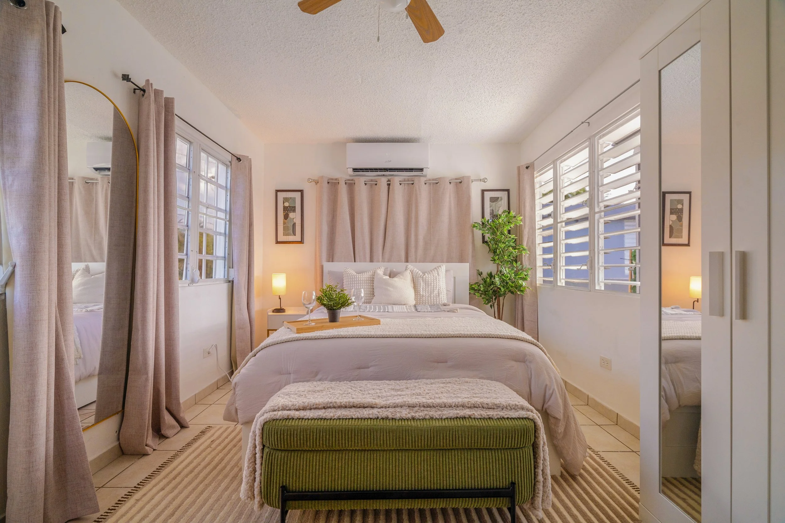 A cozy bedroom with a white bed dressed in beige and white linens, topped with decorative pillows, and flanked by green plants. The room features large windows with louvered panes, beige curtains, framed abstract artwork, and a ceiling fan. The overall decor is light and airy, with neutral tones and warm lighting.