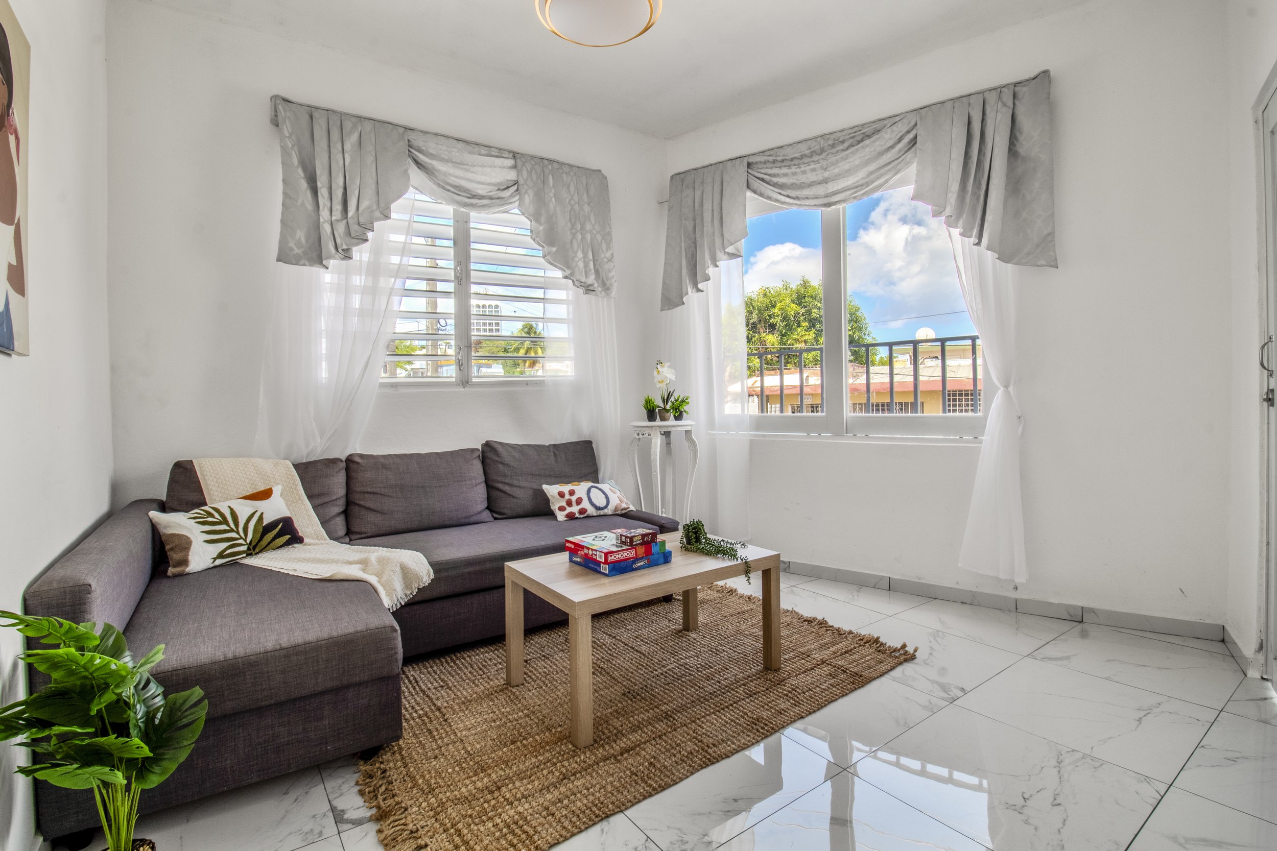 Living room with white walls, large windows with sheer white curtains and decorative gray valances, gray sofa with patterned throw pillows, small wooden coffee table with board games, potted plant with green leaves, beige rug, tiled white floor, and bright outdoor scenery visible through the windows.