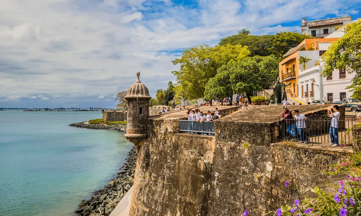 People gather along a historic stone fortress overlooking a body of water with a cloudy sky above.