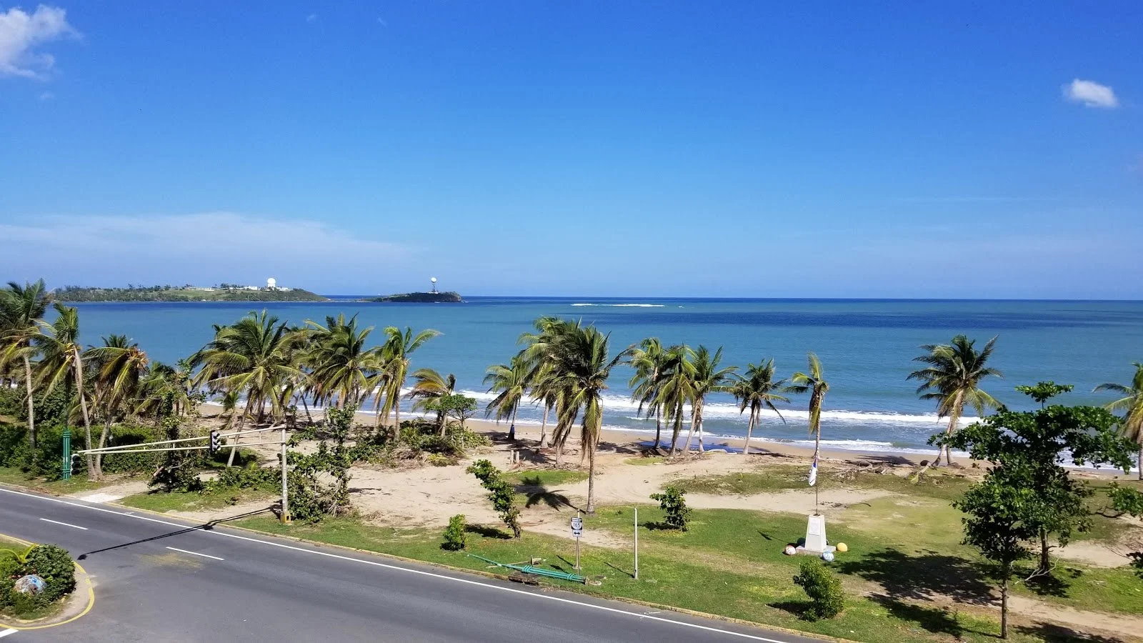 A scenic view of a tropical beach with palm trees, blue ocean water, and a clear sky, with land and radar towers in the distance.