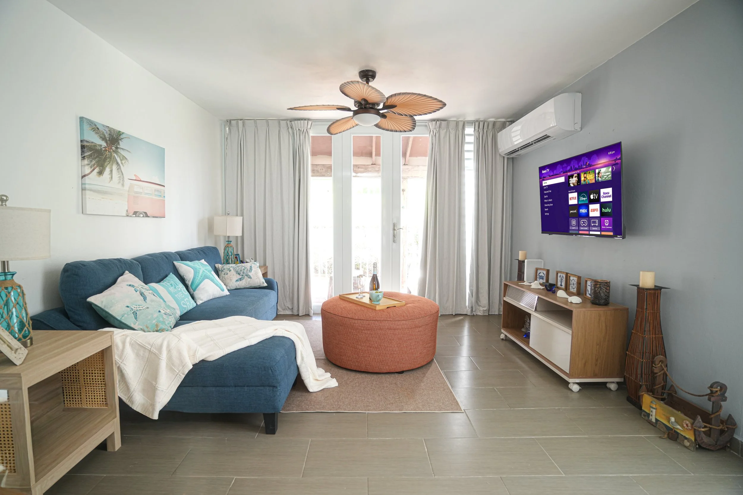 Living room with a blue sofa, throw pillows, a round orange ottoman, a framed palm tree picture, a TV mounted on the wall, and a sliding glass door with white curtains.