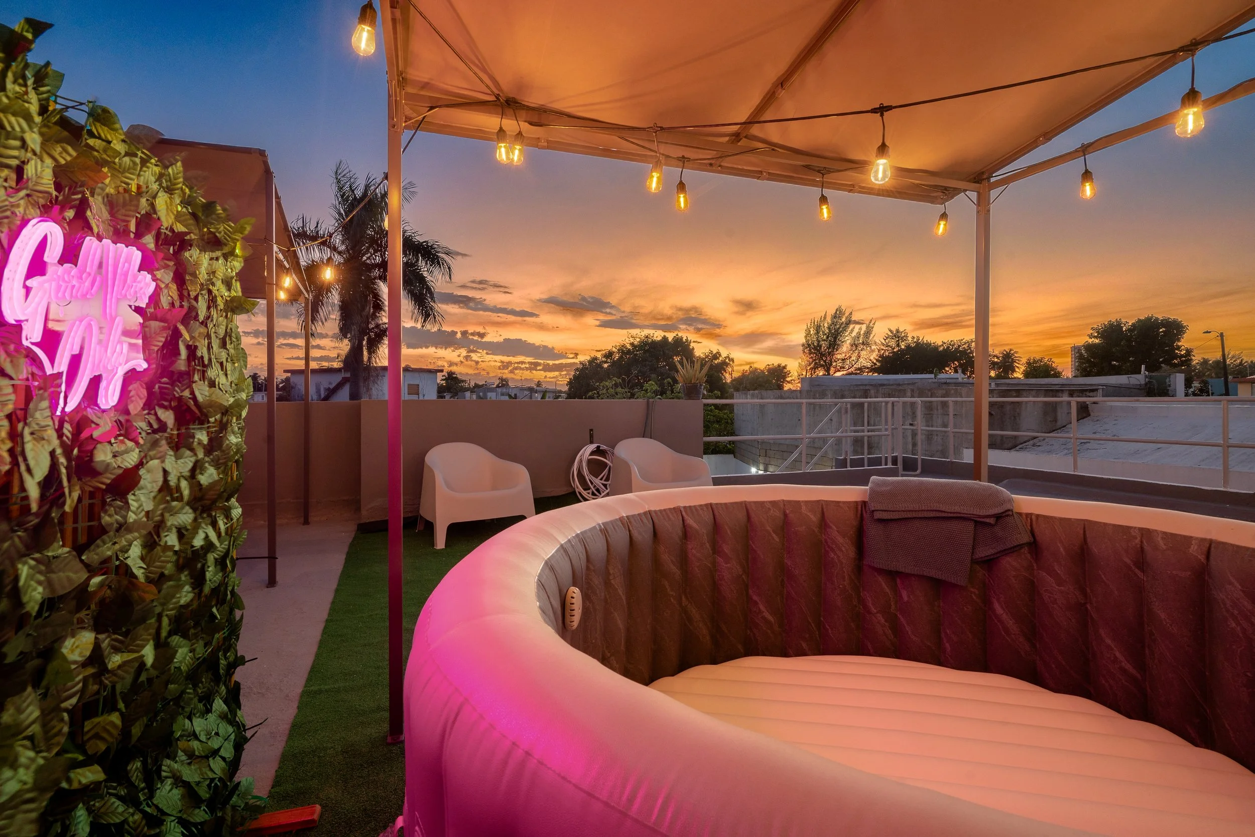 Outdoor rooftop patio at sunset with string lights, green wall, white chairs, and beige curved sofa, overlooking the sky and trees.