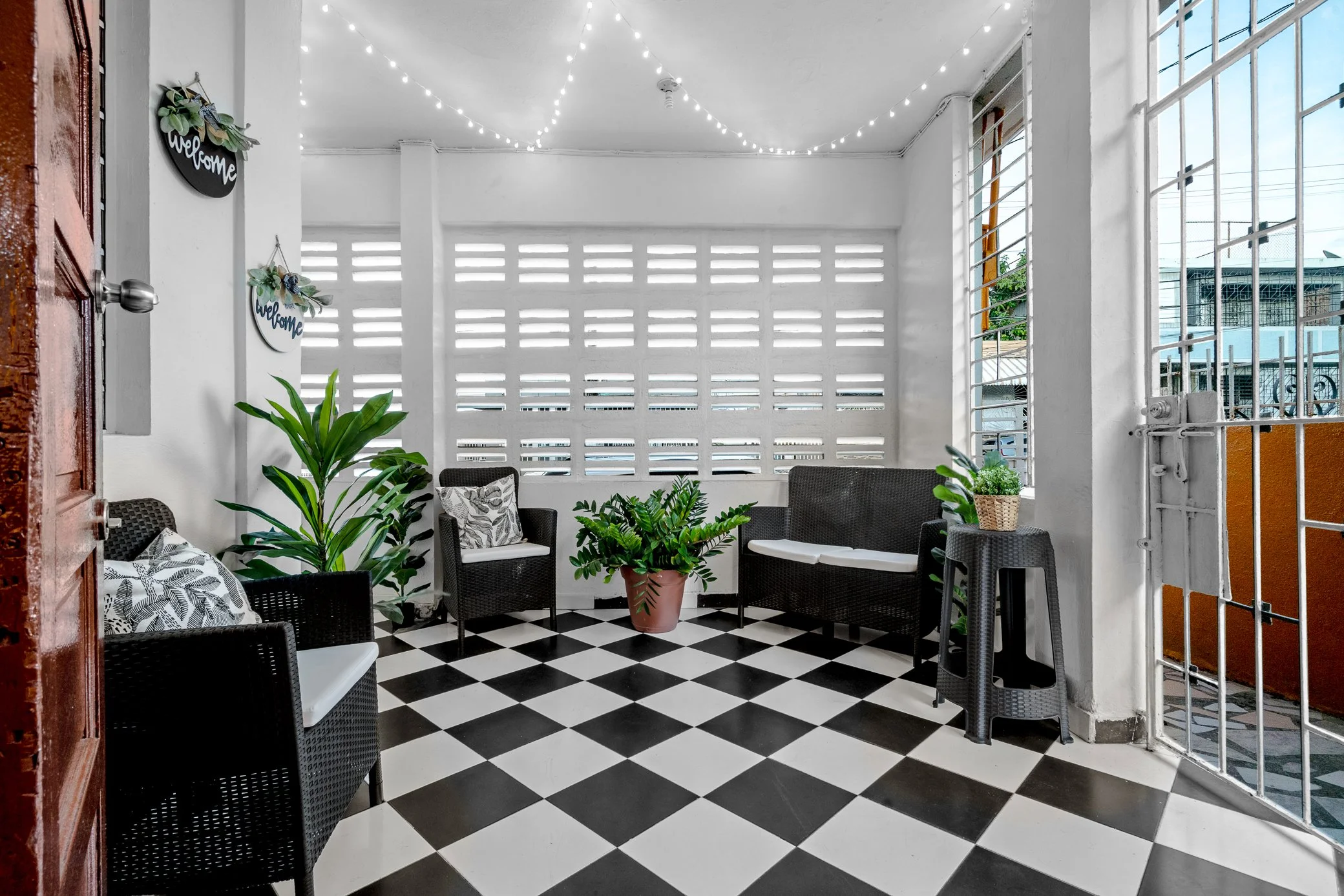 A covered outdoor seating area with black wicker chairs, white and black checkered floor, potted plants, and string lights on the ceiling.