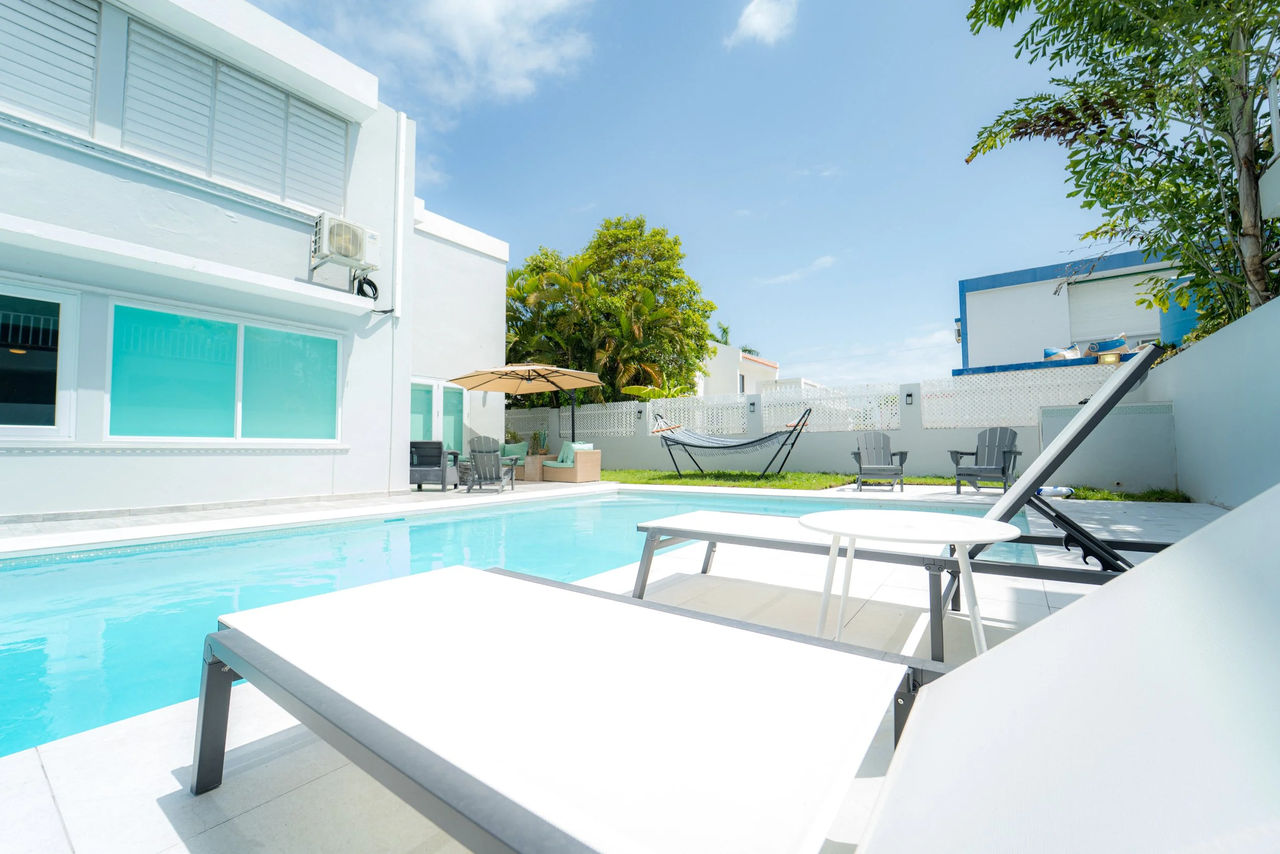 A modern residential backyard with a swimming pool, outdoor seating area with chairs and tables, umbrellas, hammock, and lush green trees under a clear blue sky.
