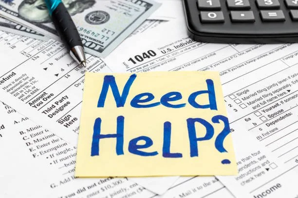 A yellow sticky note with blue handwritten text reading 'Need Help?' on a cluttered desk with a pen, U.S. tax forms, and a calculator.
