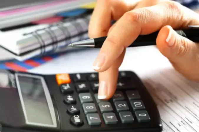 A person pressing calculator keys with a pen in hand, on a desk with papers and notebooks.