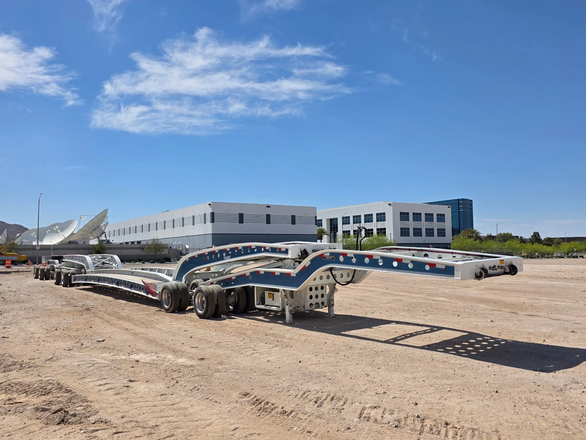 Empty flatbed trailer parked on dirt ground with a building and satellite dishes in the background, under a blue sky with some clouds.