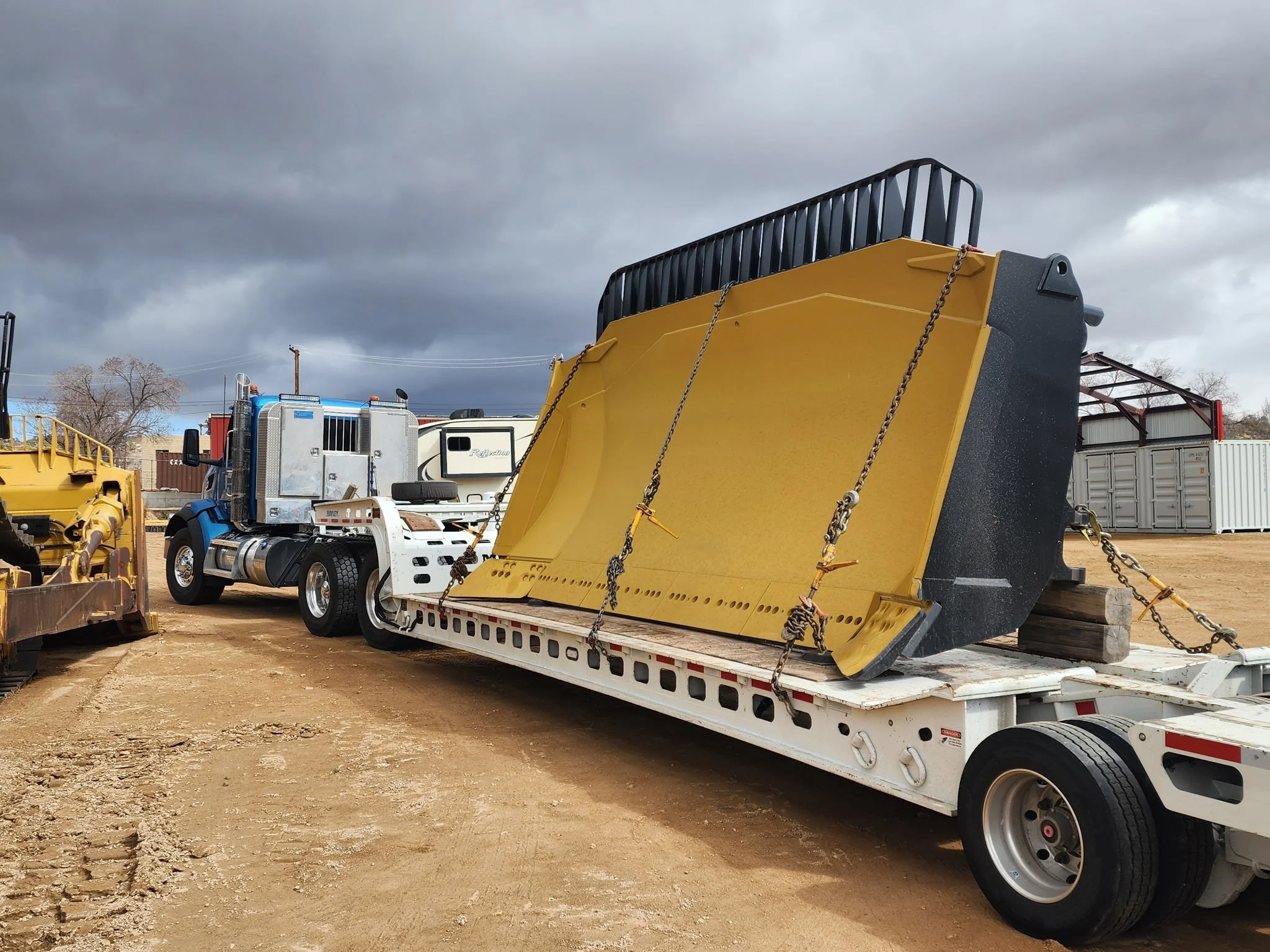 A yellow snow plow blade secured on a flatbed trailer with chains, parked outdoors on dirt ground, with other trucks, storage containers, and overcast sky in the background.