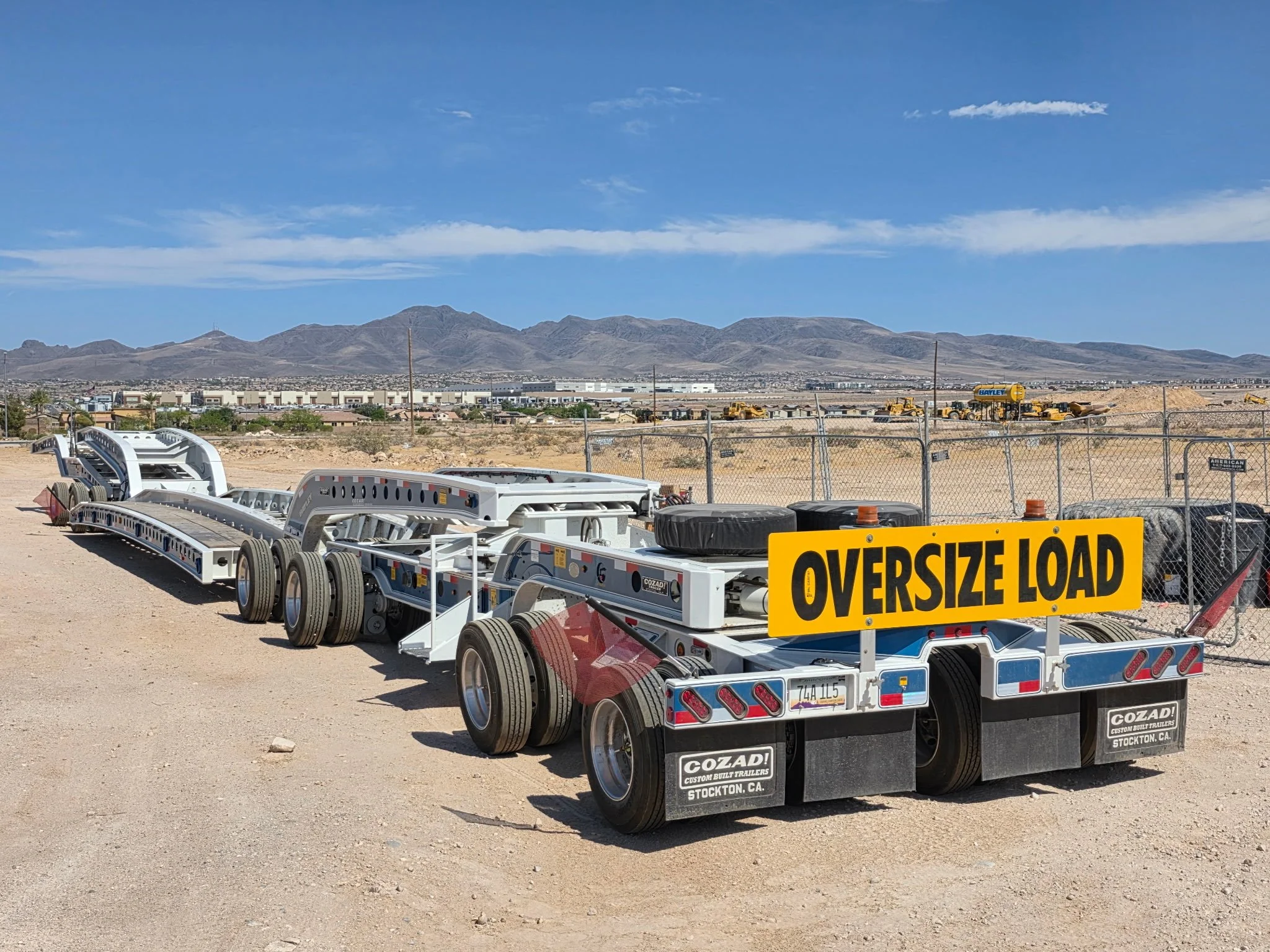 A parking lot with multiple trailer trucks, including one with a sign that reads American Oversize Load. The background features a desert landscape with mountains and a partly cloudy sky.