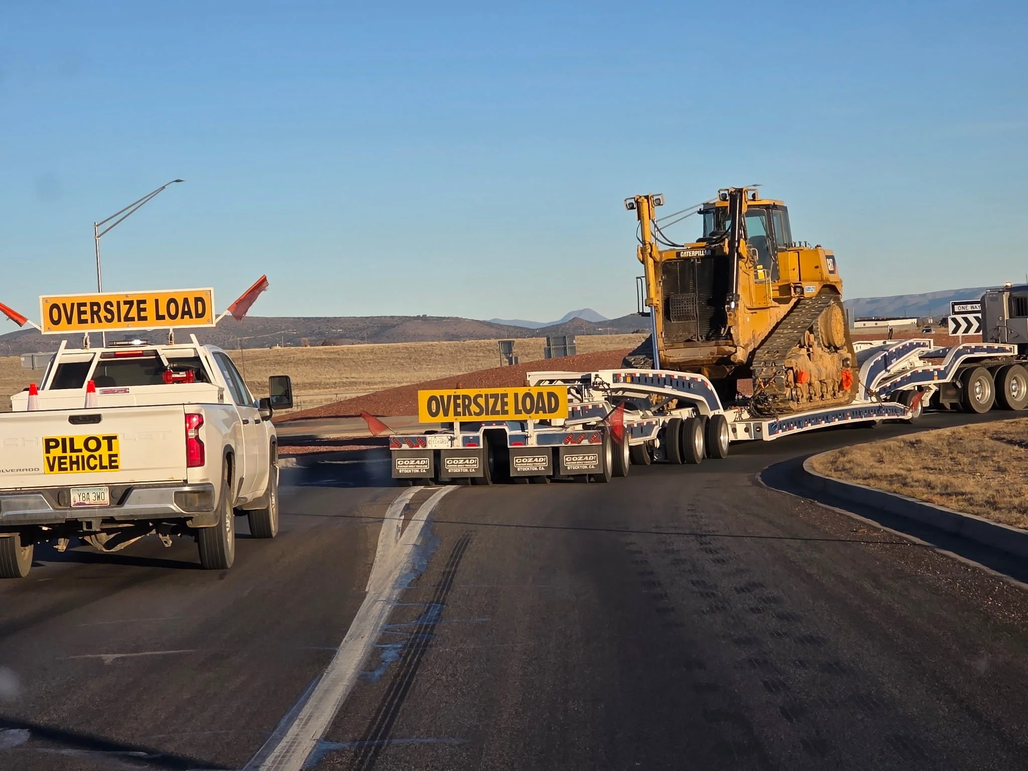 A construction zone on a highway with a large yellow Caterpillar excavator on a flatbed trailer, signs indicating an oversized load and a pilot vehicle, and orange traffic cones, with a distant mountain range and clear blue sky in the background.