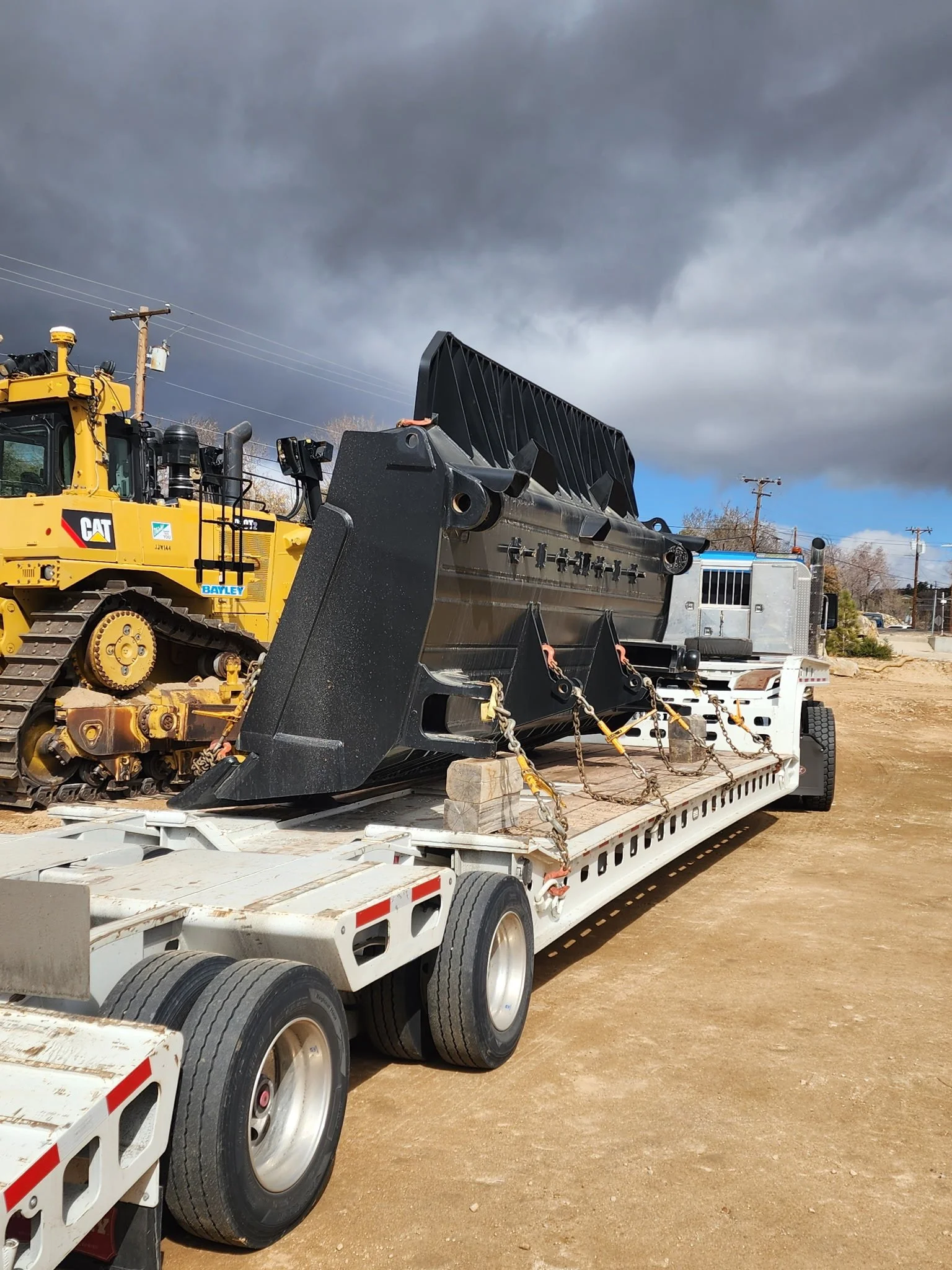 A flatbed truck carrying construction equipment, including a yellow Caterpillar bulldozer and a large black attachment, parked on a dirt lot under a cloudy sky.