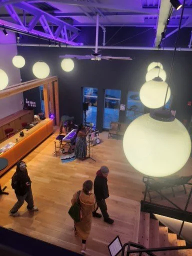 Indoor event space with large hanging spherical lights, a music performance area with a keyboard and microphone, and several people walking around.