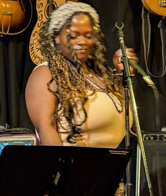 A woman with long, gray-dyed dreadlocks smiling while holding a microphone on stage, surrounded by musical instruments.