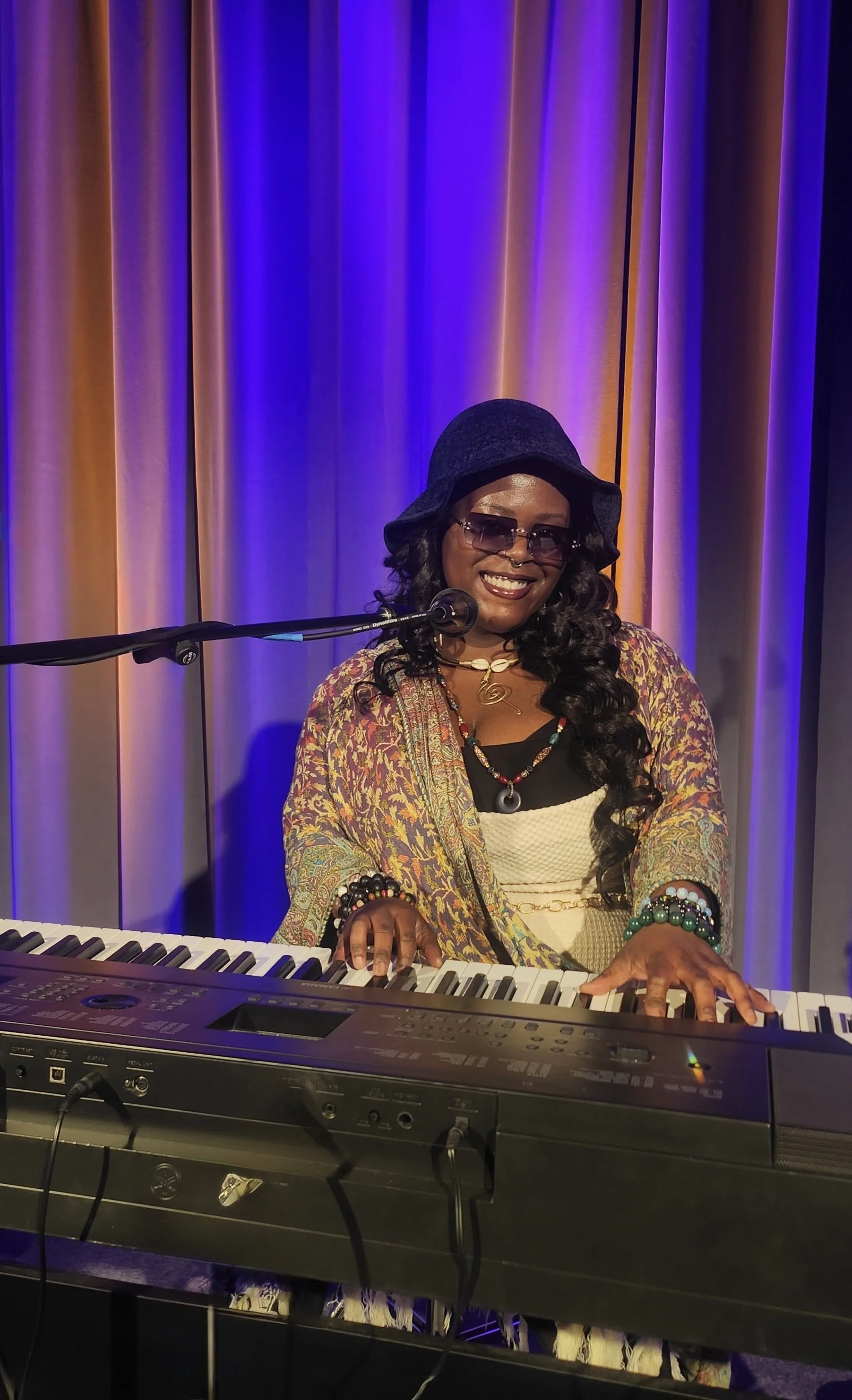 A woman with dark curly hair, wearing sunglasses, a black hat, colorful jewelry, and a paisley shawl, playing a keyboard on stage with purple and orange curtains in the background.