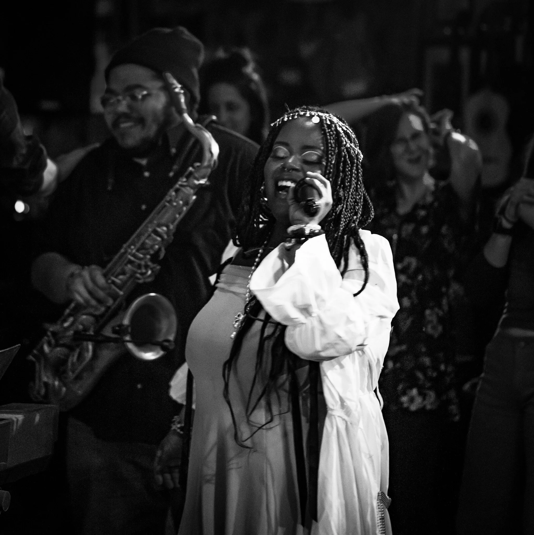 A black and white photo of a group of people at a social gathering, with one woman singing or speaking into a microphone and others possibly playing instruments or engaged in conversation, in a dimly lit setting.